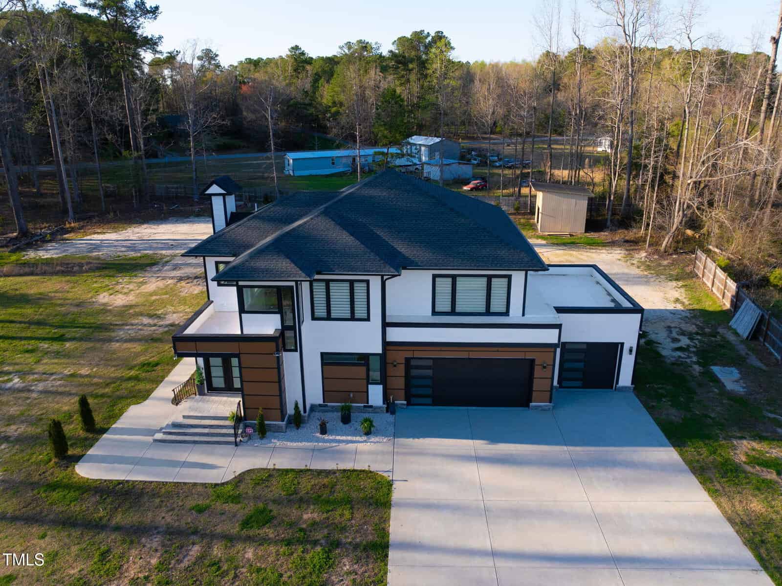 A modern two-story house with a flat roof, large windows, and a three-car garage sits on a spacious lot with a concrete driveway, surrounded by grass, trees, and neighboring structures in the background.