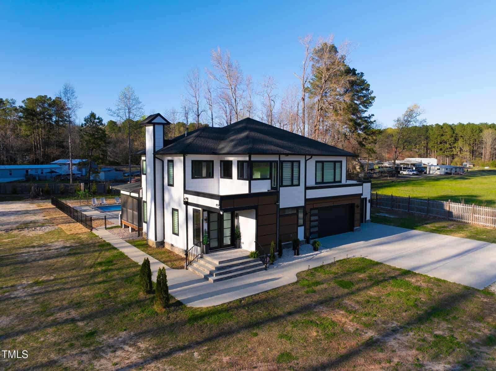 A modern two-story house with black and white exterior, large windows, an attached three-car garage, and concrete driveway; surrounded by grass, trees, and neighboring homes in a rural area.