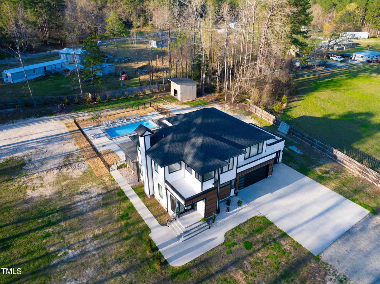 Aerial view of a modern two-story house with a black roof, attached garage, fenced backyard, and rectangular swimming pool; surrounded by grass, trees, a shed, and neighboring properties.