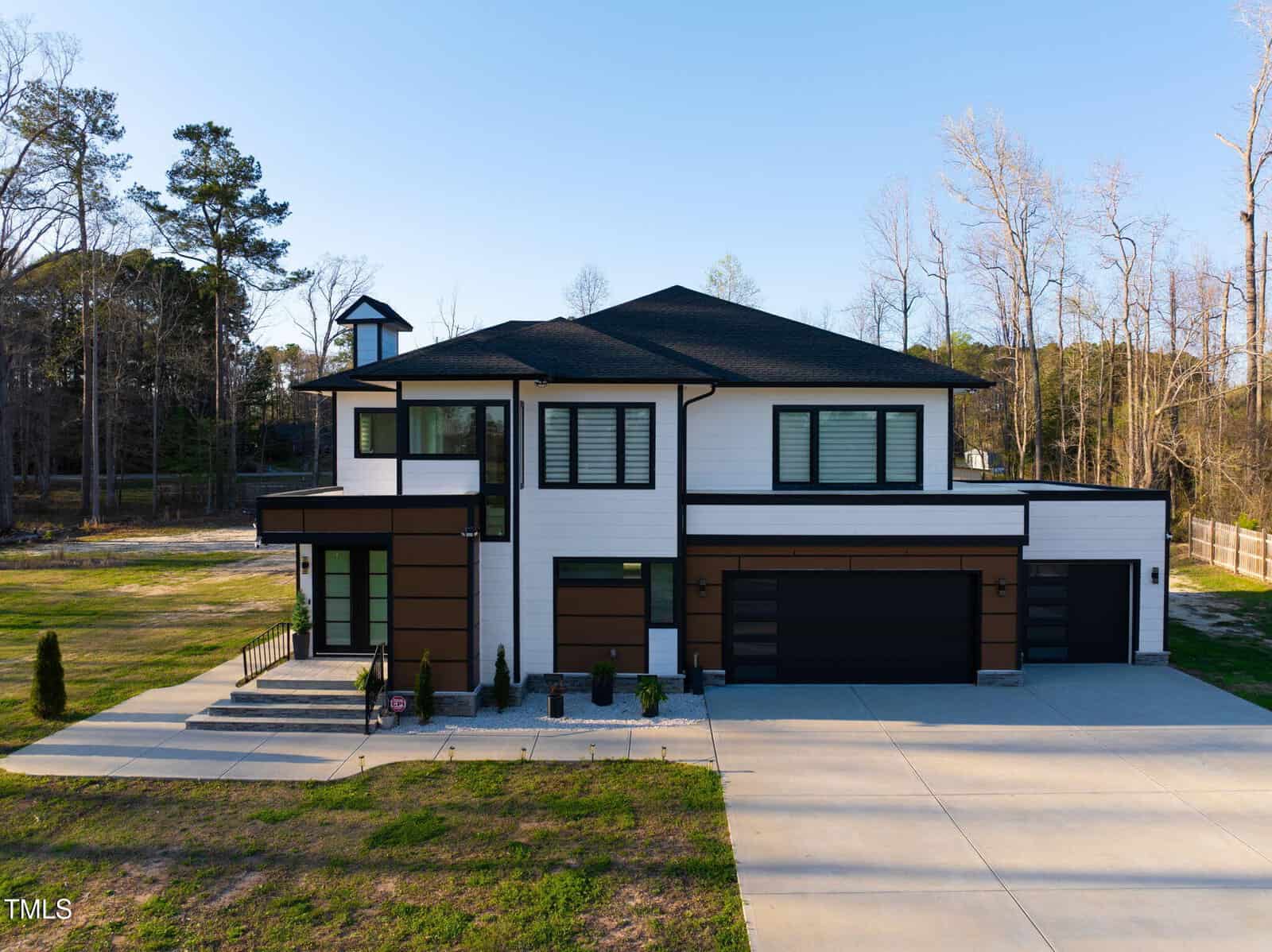 A modern two-story house with white and brown exterior panels, large black-trimmed windows, a three-car garage, and a wide driveway. The property is surrounded by trees and has minimal landscaping.