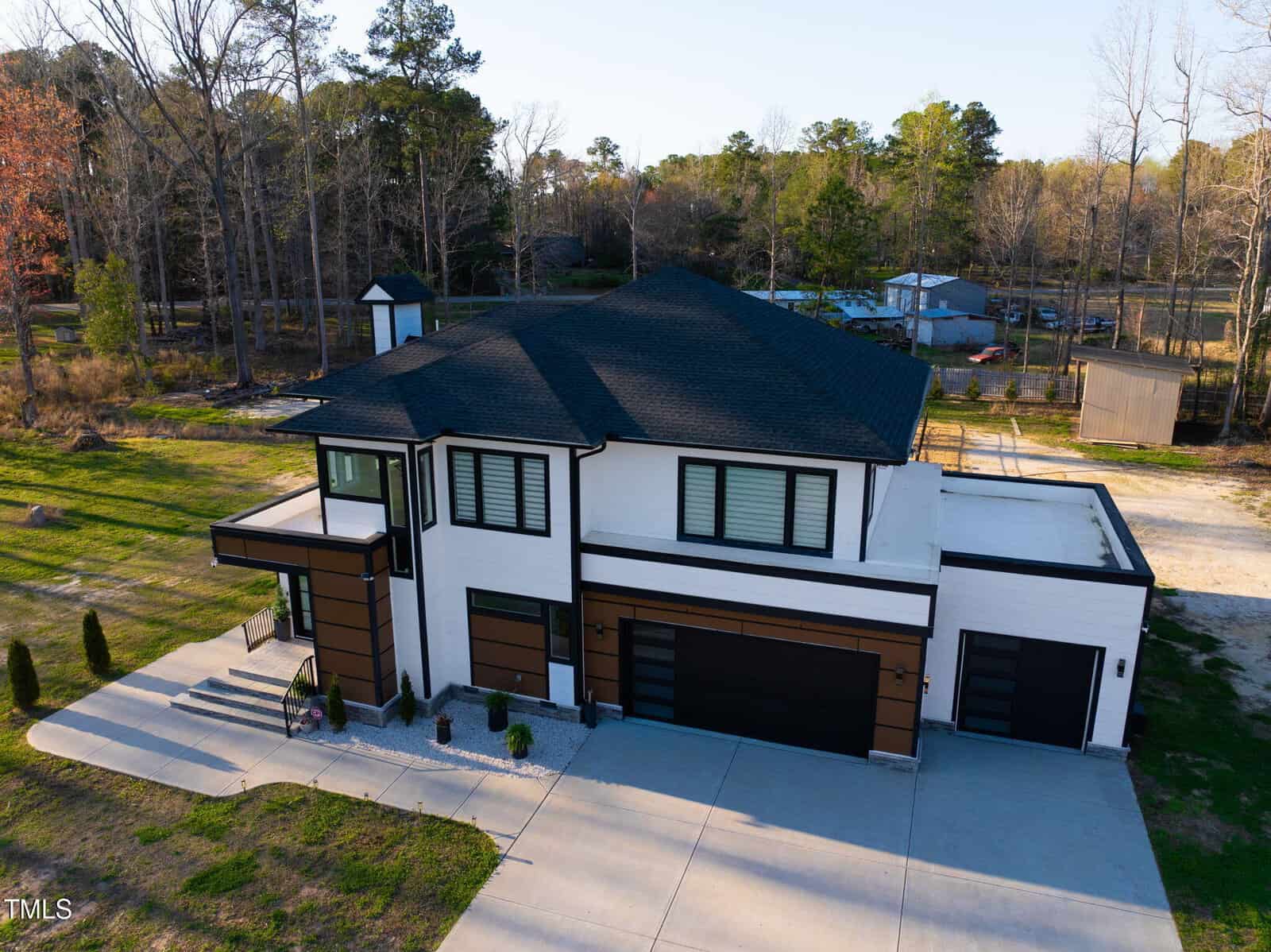 Modern two-story house with white and brown exterior, large windows, black roof, and three-car garage. Surrounded by driveway, sparse landscaping, and trees in the background.
