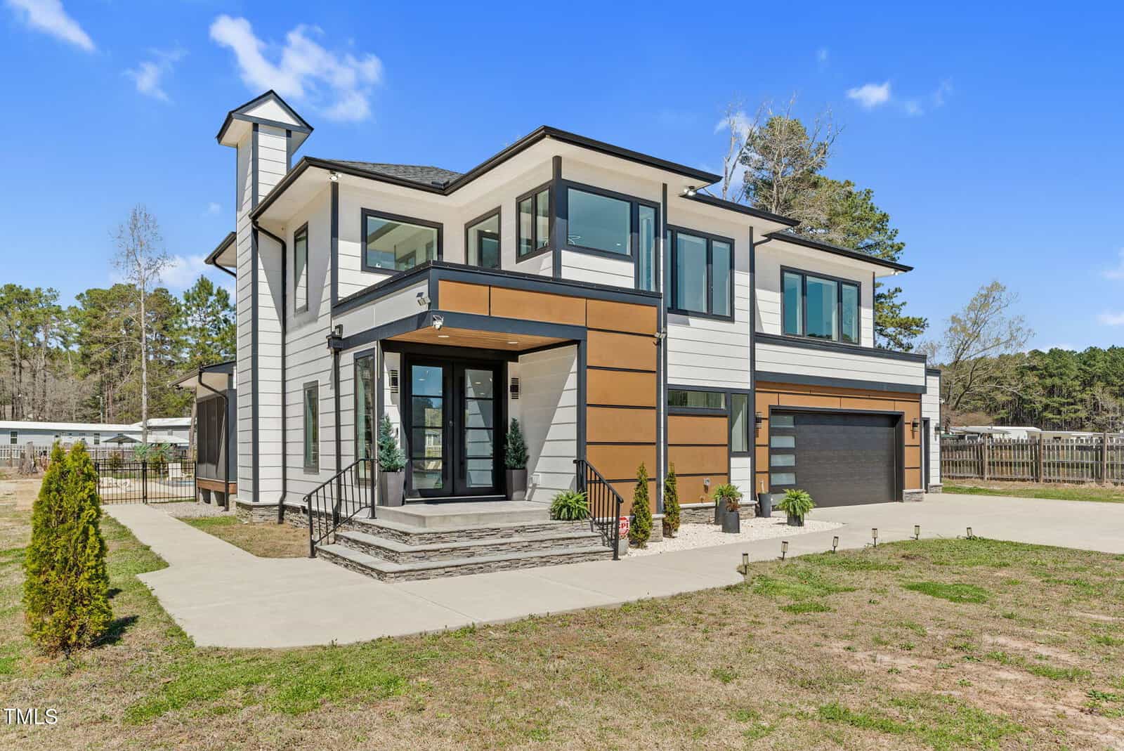 A modern two-story house with large windows, white and brown exterior panels, a black roof, and a double garage. The front entrance has steps, potted plants, and the surrounding area has sparse grass and a paved driveway.