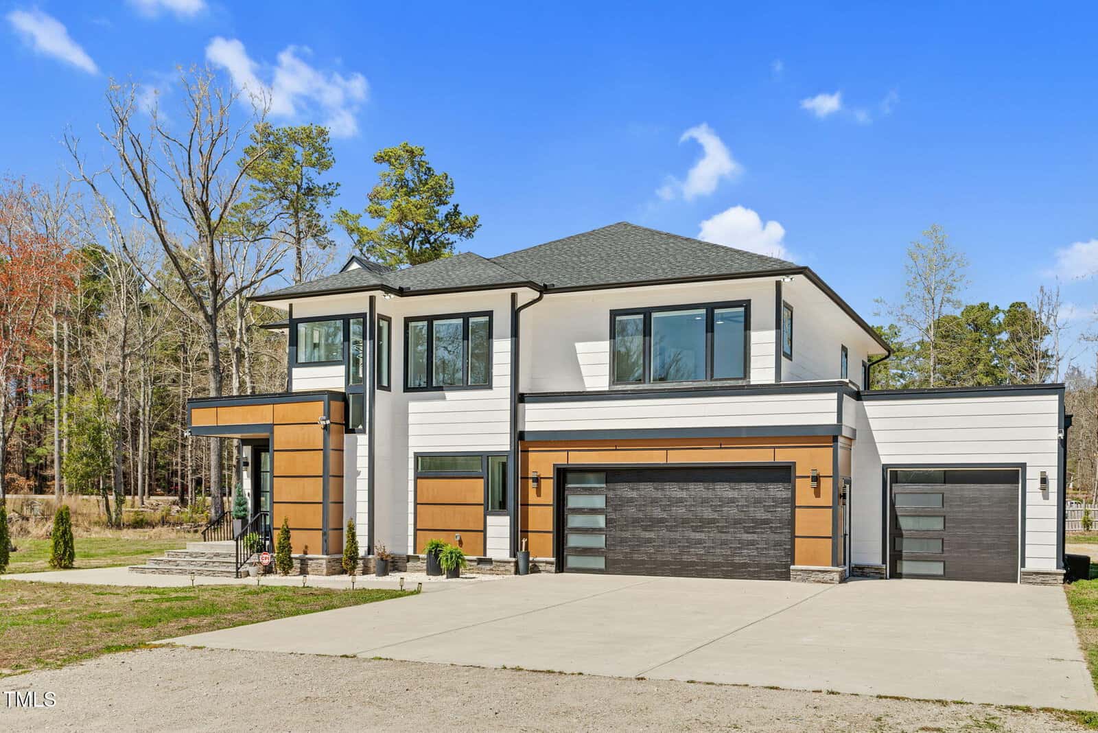 Modern two-story house with white and wood-paneled exterior, black trim, large windows, and two garages. The driveway is concrete, and the surrounding area includes trees and a clear blue sky.