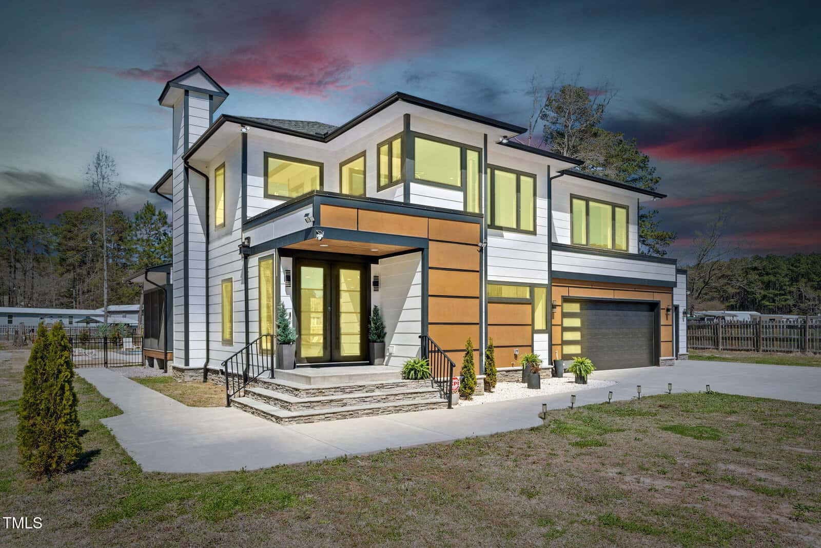 Modern two-story house with large yellow-tinted windows, white and wood-accent siding, flat roof, and attached garage. The home sits on a concrete driveway with minimal landscaping, at dusk with a colorful sky in the background.