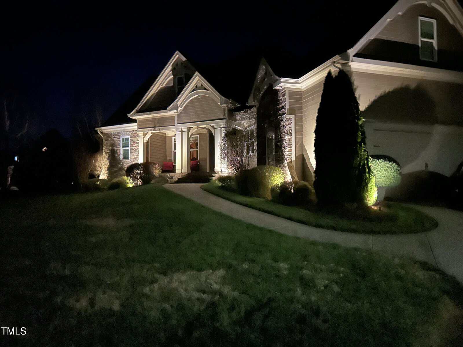 A well-lit house at night with exterior lights highlighting its facade. The house has a porch and a curved walkway leading to the entrance. Shrubs and trees line the path. The front lawn is visible in low light.