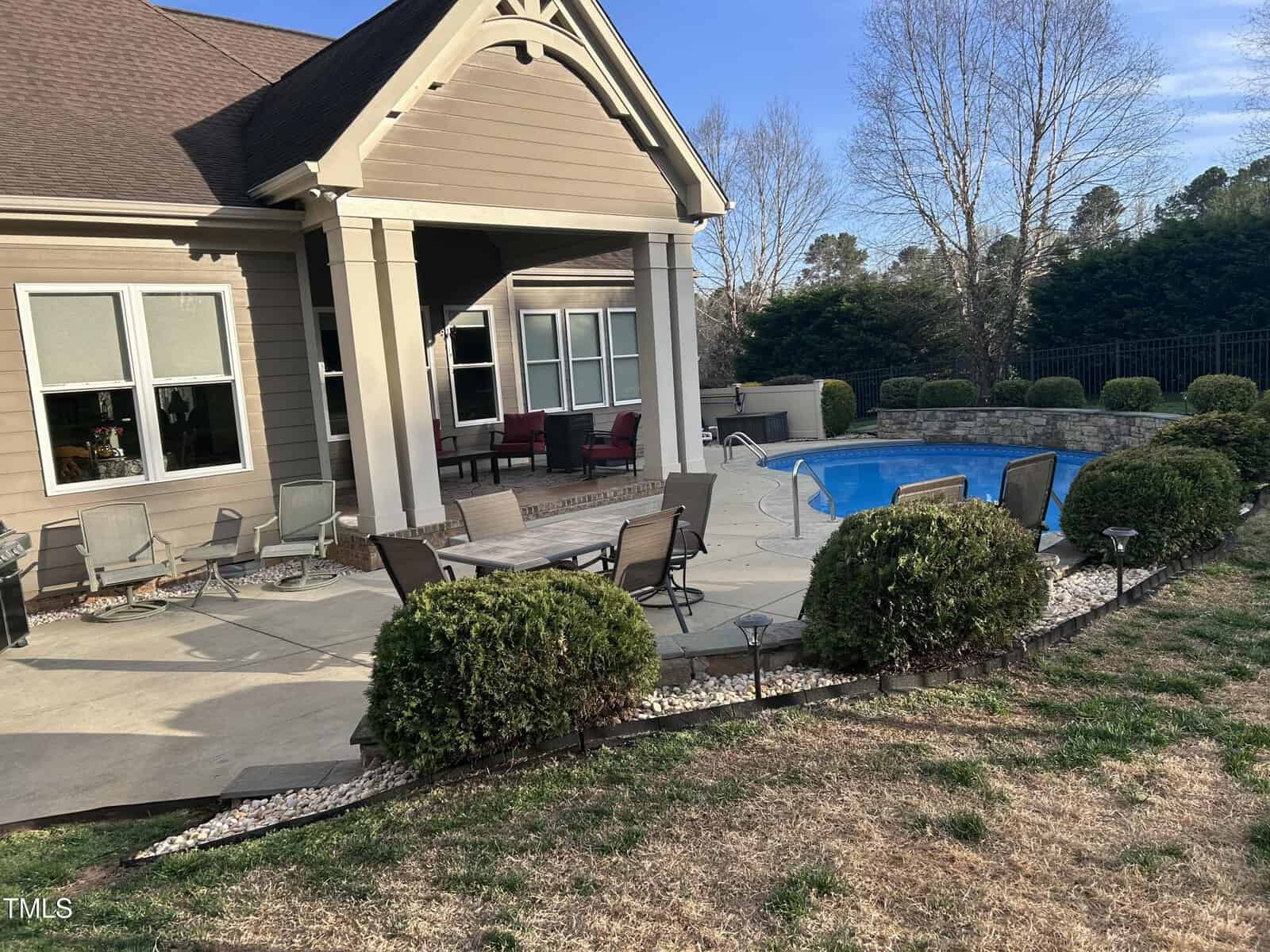 A backyard patio with a table, chairs, and umbrella set on a concrete surface. A swimming pool is nearby, surrounded by a low stone wall. The house has a covered porch with additional seating. Bushes and trees line the yard.