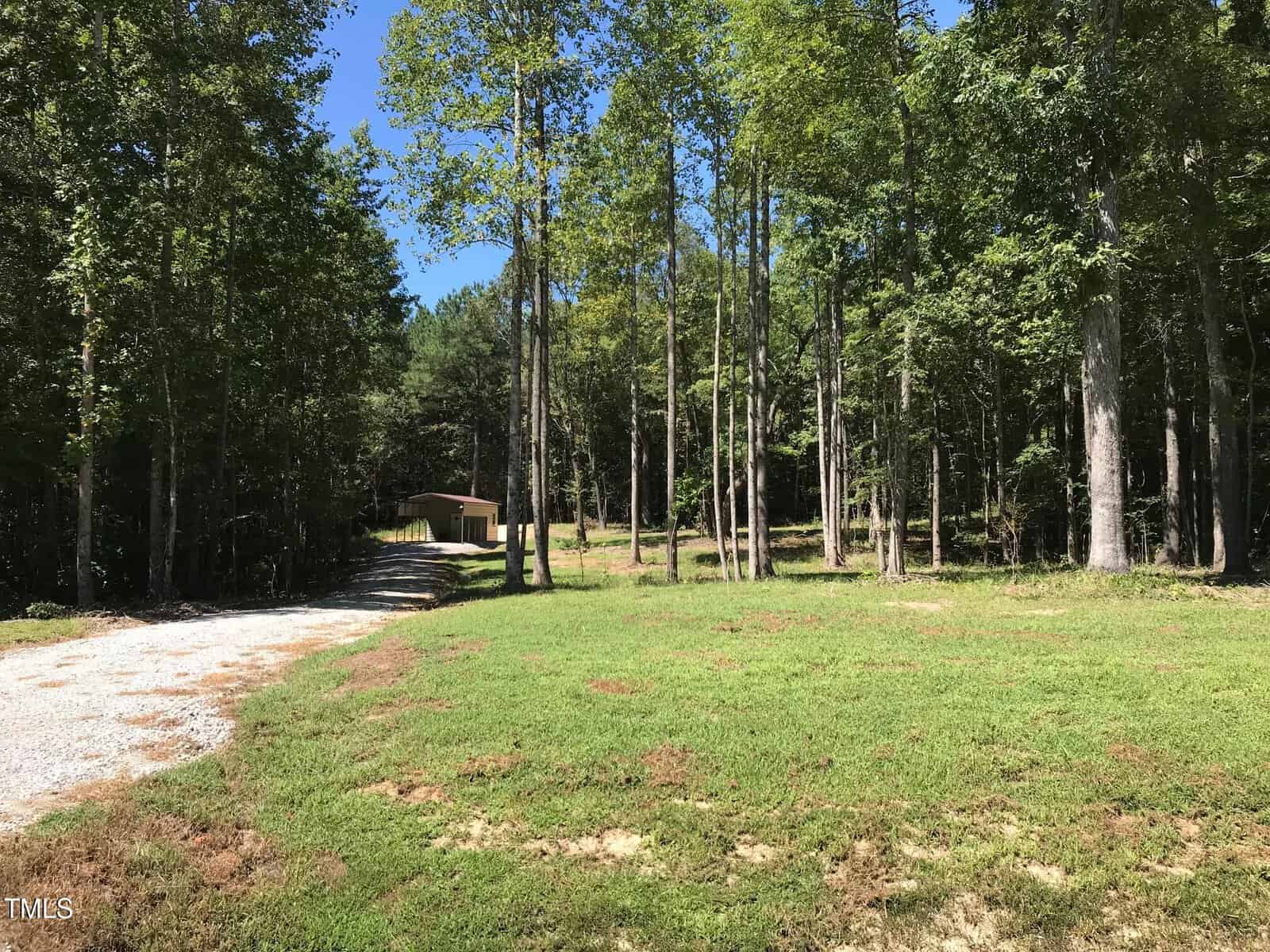 A rural scene with a gravel driveway leading to a small house surrounded by tall trees. The foreground features open grassy areas, and there is a clear blue sky above.