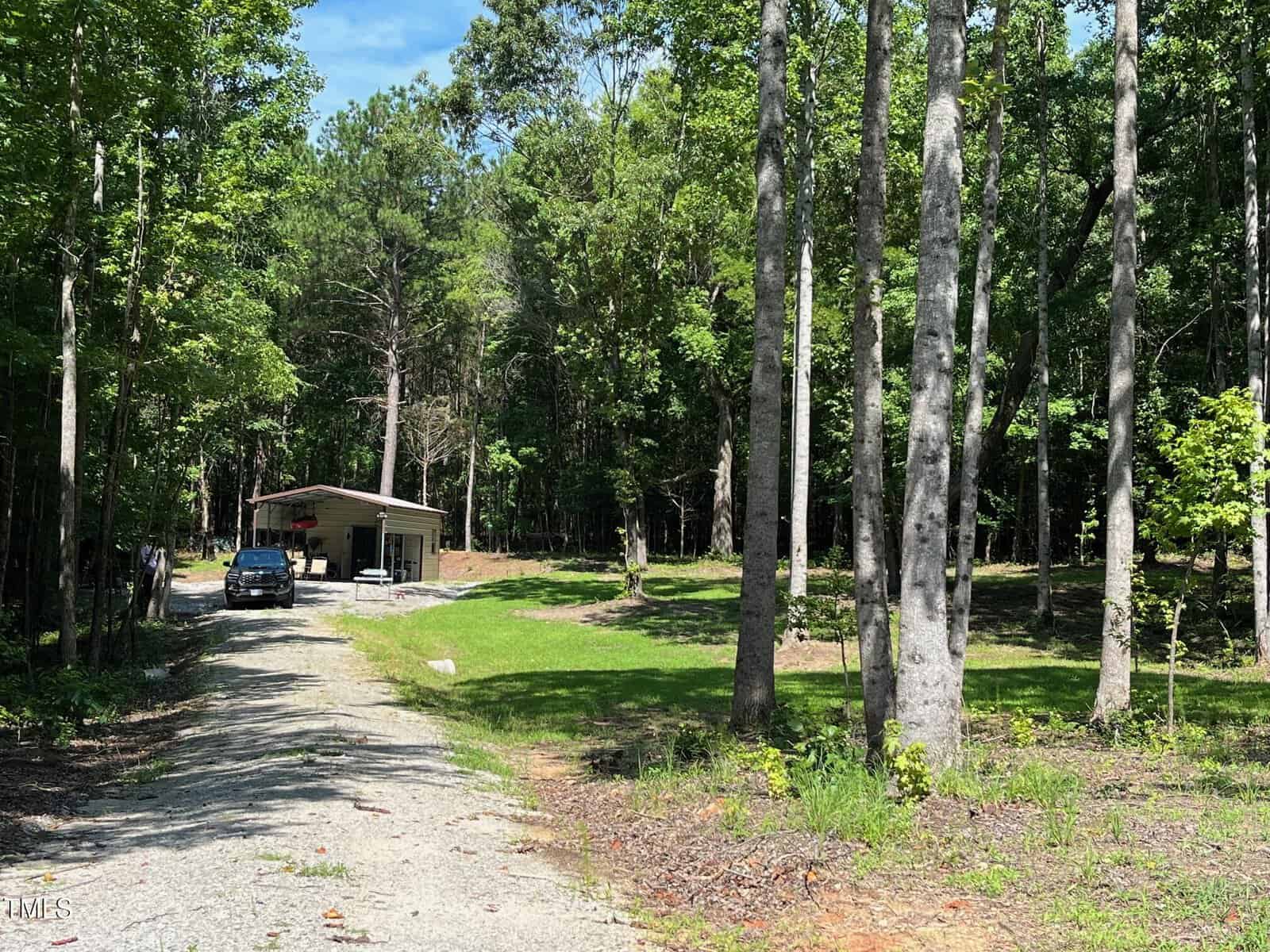 A gravel road leads to a small house with a car parked in front, surrounded by tall trees and greenery under a clear blue sky.