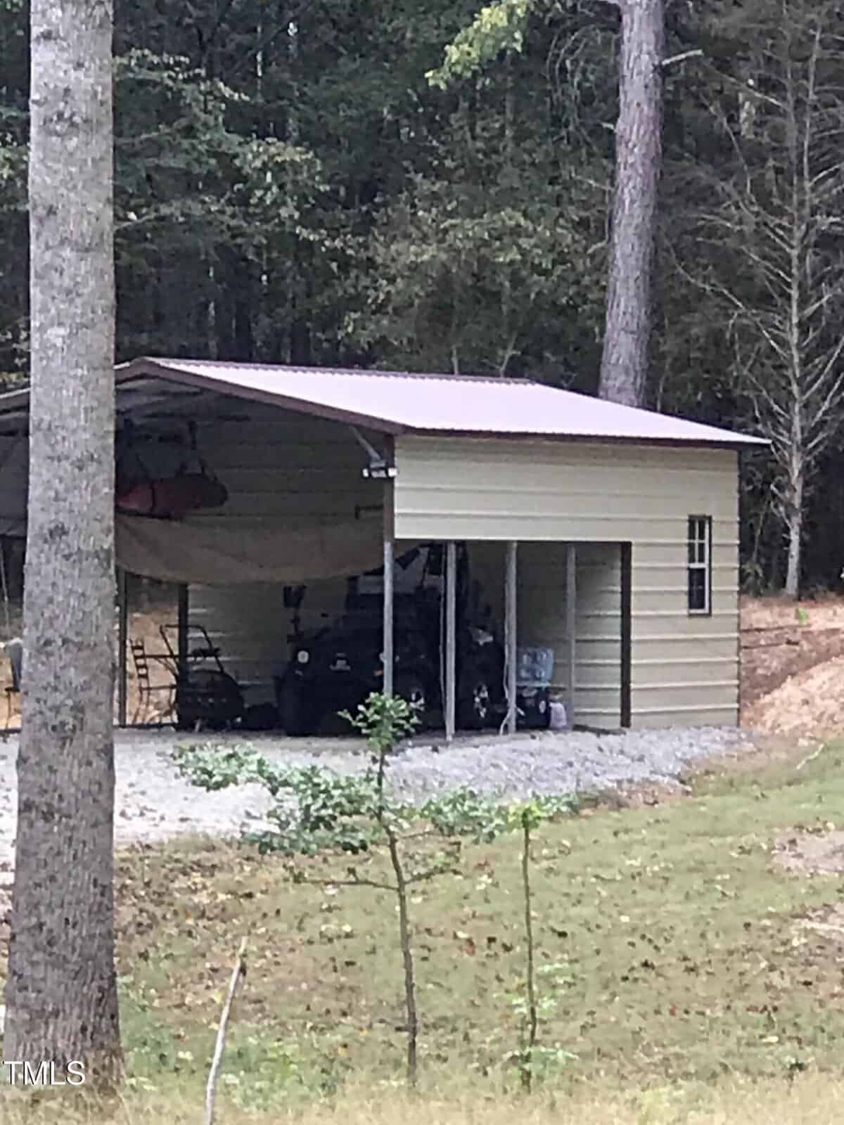 A metal carport stands in a wooded area. It houses an off-road vehicle, various tools, and equipment. The structure has a slanted roof and open sides. Trees and a grassy area with sparse underbrush surround the building.