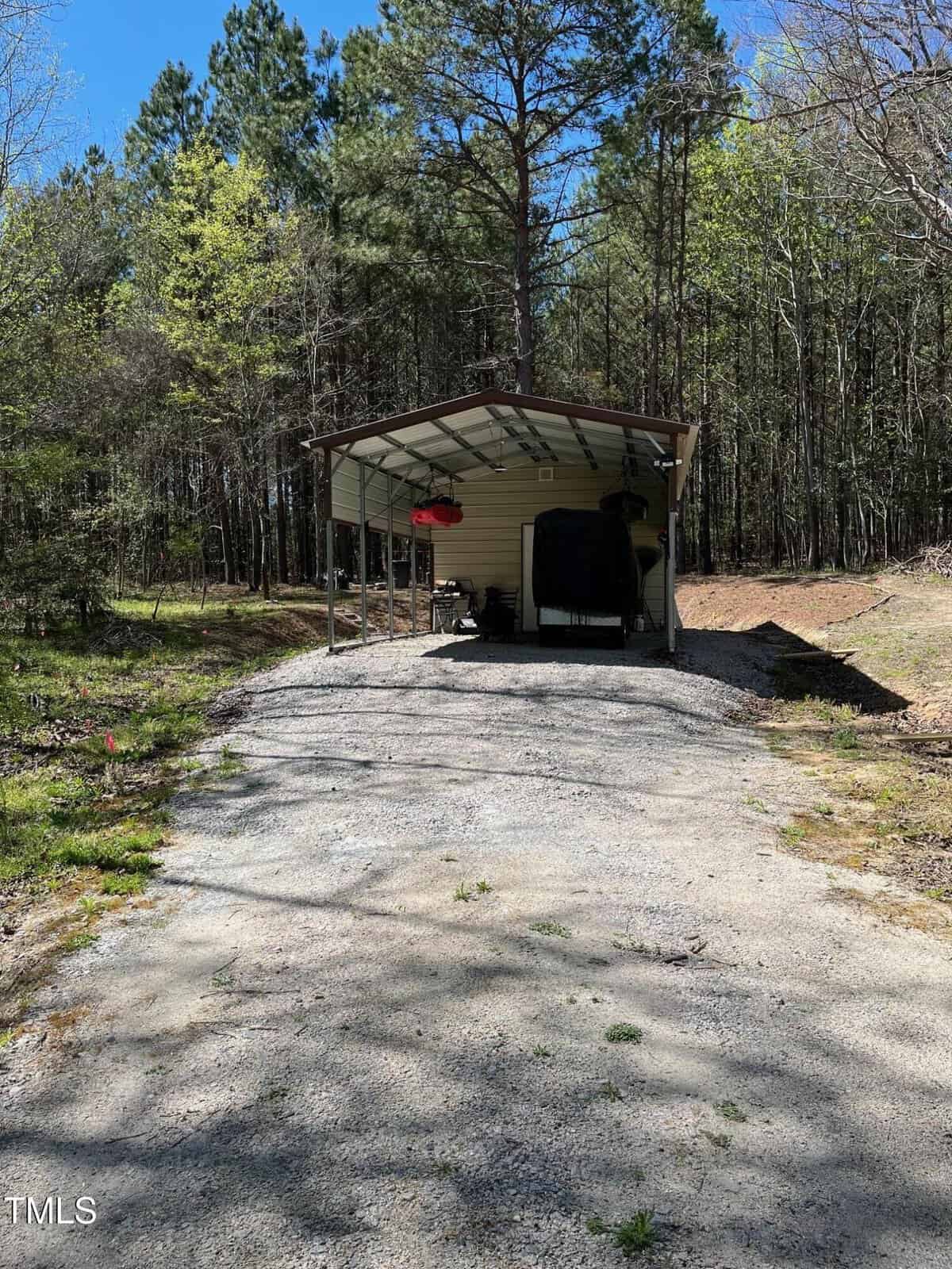 A covered shelter stands on a gravel driveway surrounded by trees. The shelter houses a motorcycle and a lawnmower. A patch of forest with green foliage is visible in the background.