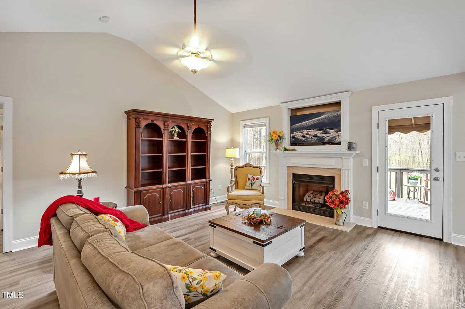 Living room with beige walls, a tan sofa, a wooden coffee table, a fireplace, a large cabinet, a chair, and a door leading outside. The room has wood flooring, a ceiling fan, and decor like flowers and paintings.