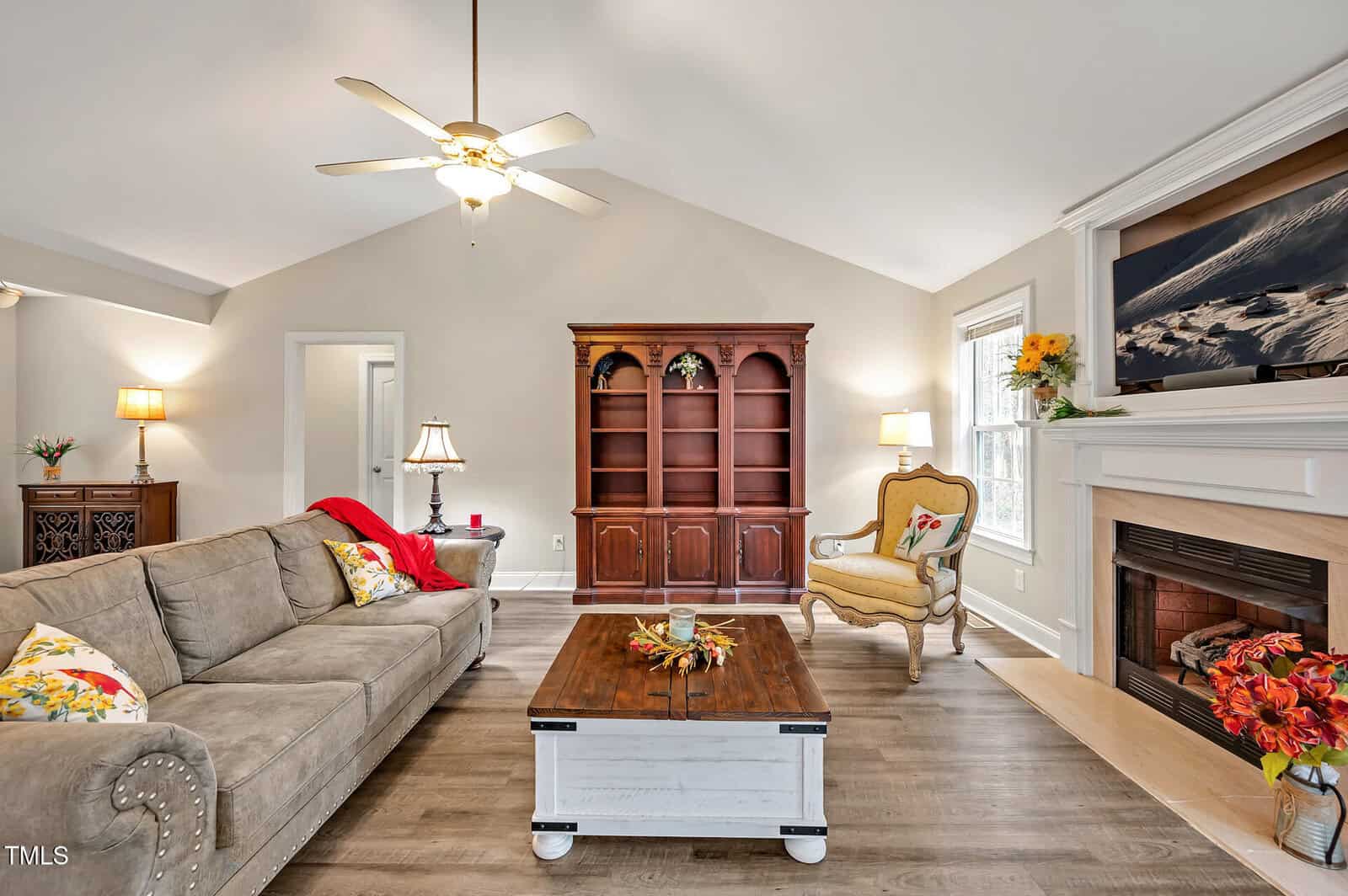 A living room with a beige couch, patterned armchair, wooden coffee table, empty wooden shelves, and a fireplace with a fall-themed mantel, under a ceiling fan with lights. A window lets in natural light.