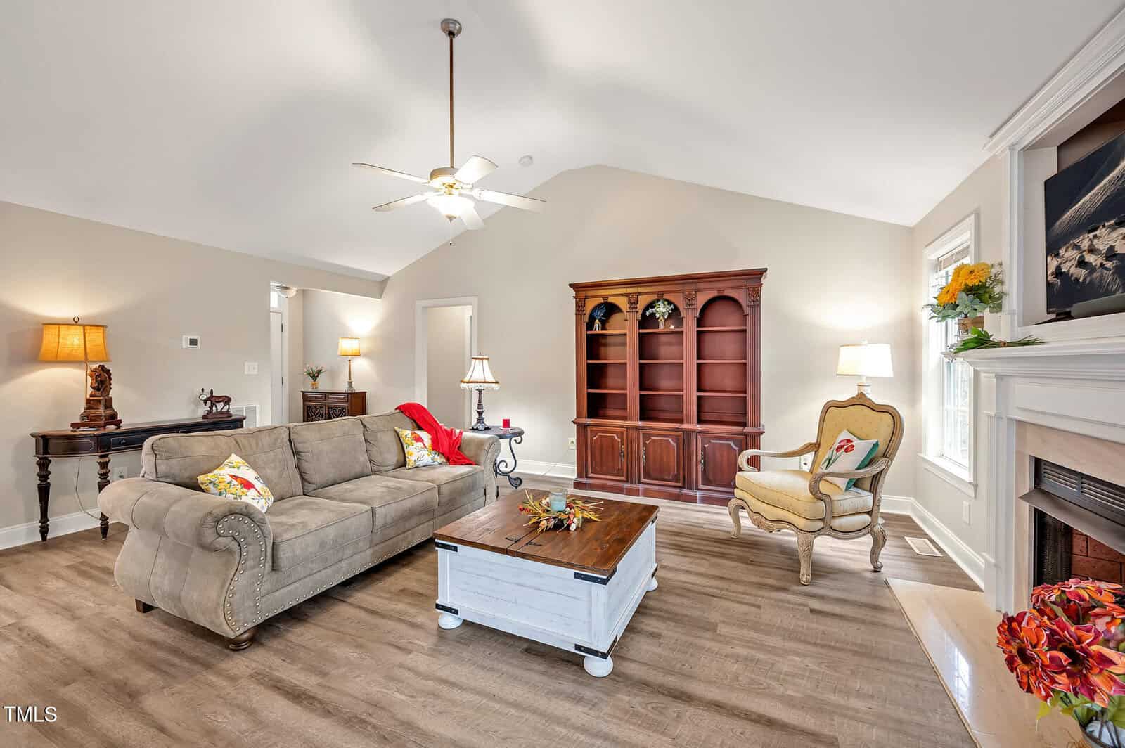 A living room with a beige sofa, wooden coffee table, armchair, and empty wooden bookshelf. A lit lamp sits on a small table, and a fireplace with a mantel is decorated with flowers. The walls are light-colored.