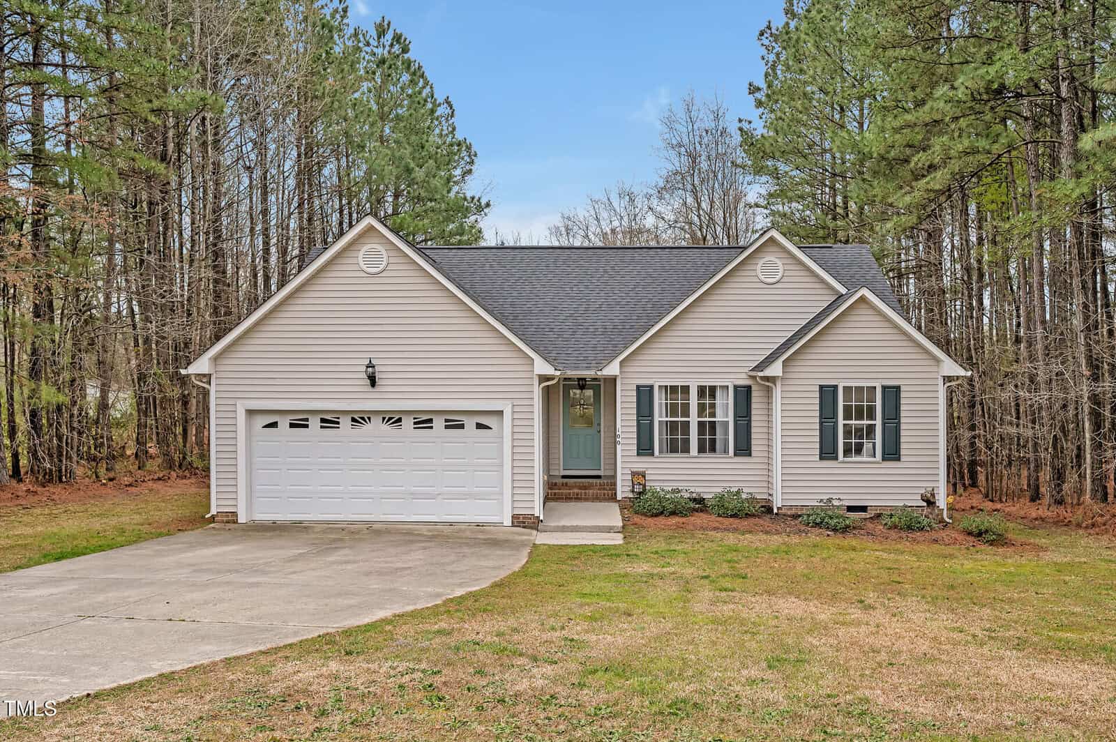 A single-story house with beige siding, dark shutters, a two-car garage, and a concrete driveway, set in front of a wooded area with pine trees under a blue sky.