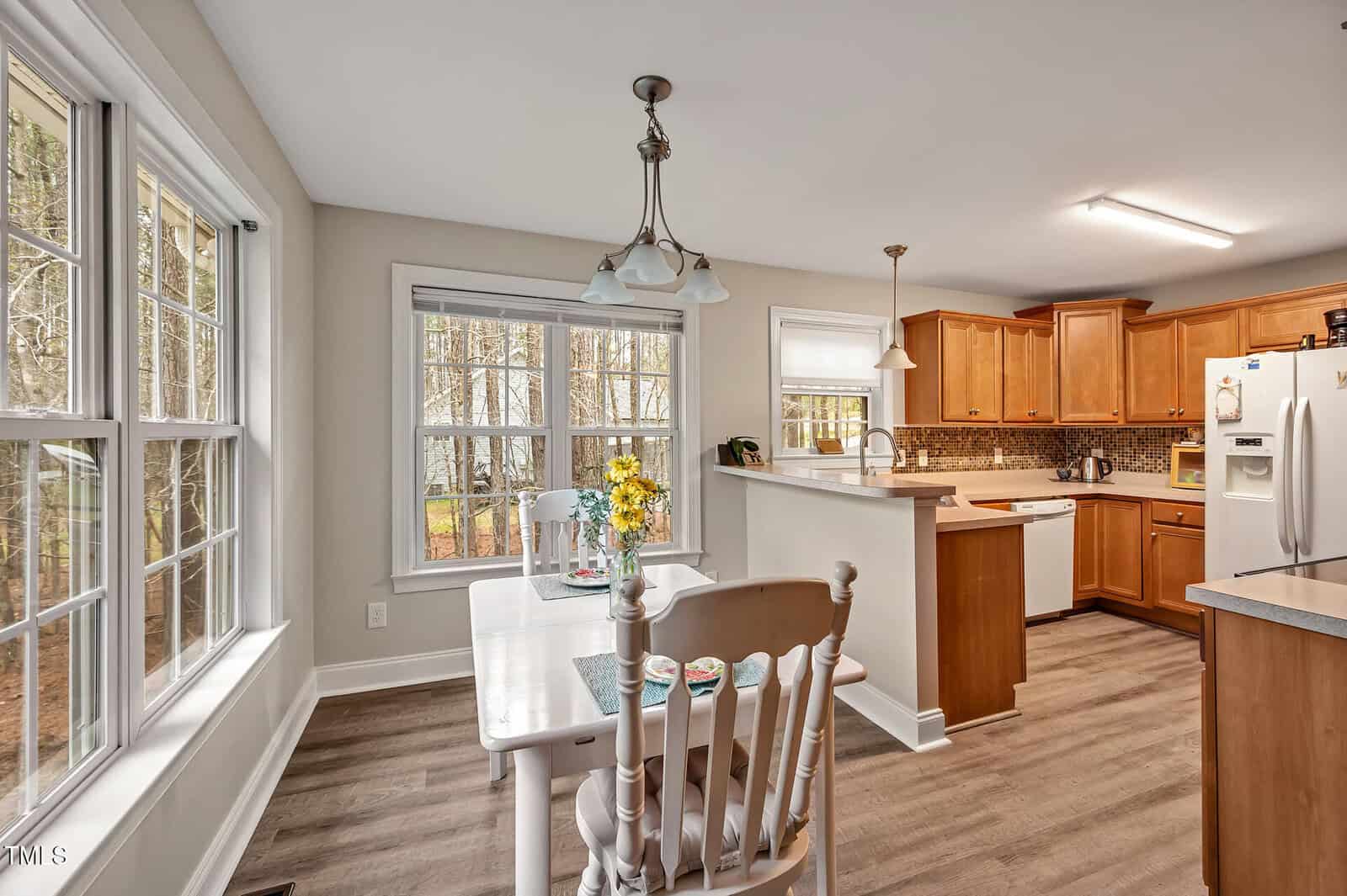 A kitchen and dining area with wood cabinets, white appliances, a white dining table with chairs, flowers on the table, large windows, pendant light, and wood-look flooring. Trees are visible outside through the windows.