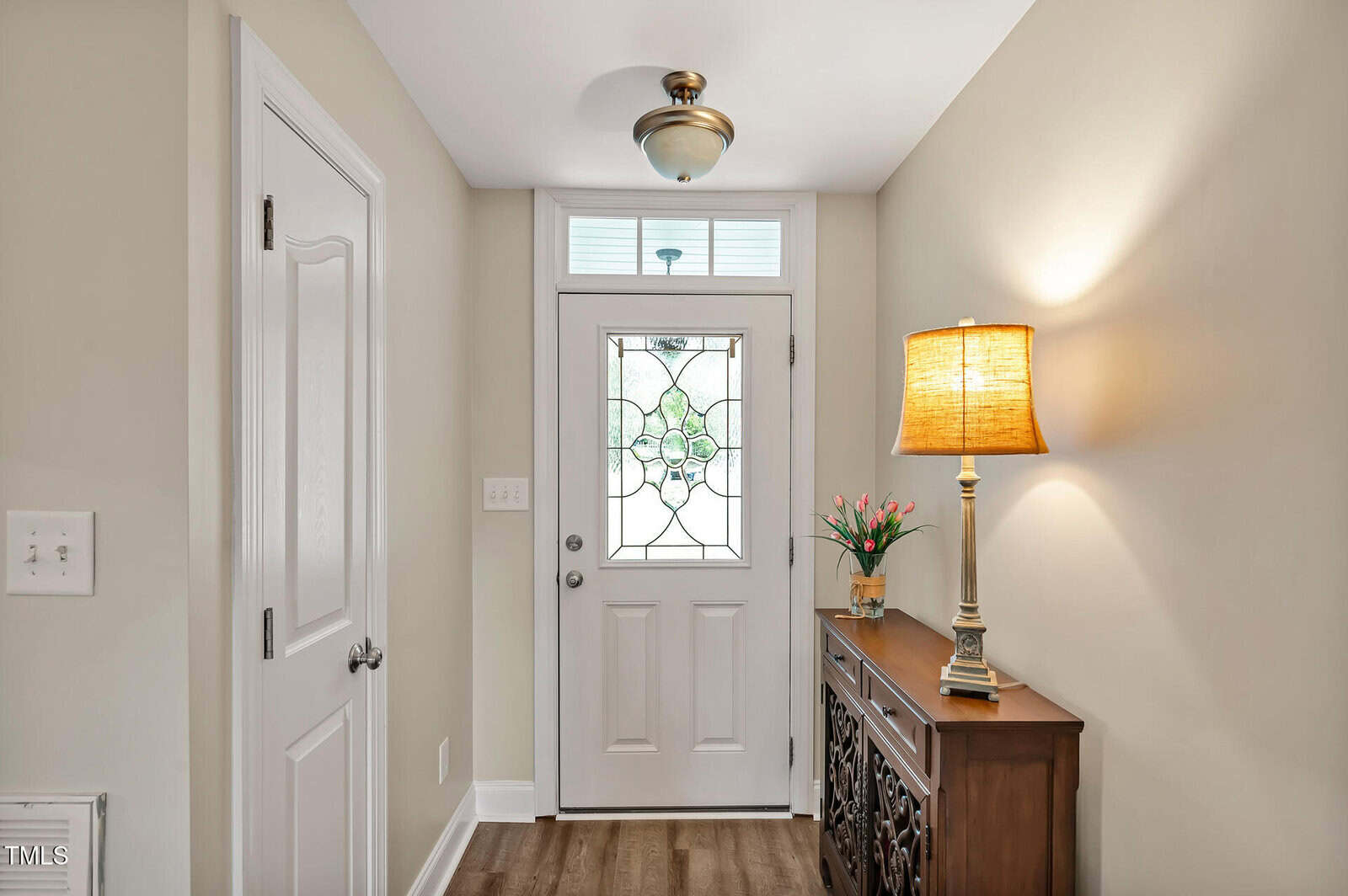 A hallway with a white front door featuring decorative glass, a small window above the door, a wooden side table with a lamp and a vase of flowers, beige walls, and wood flooring.