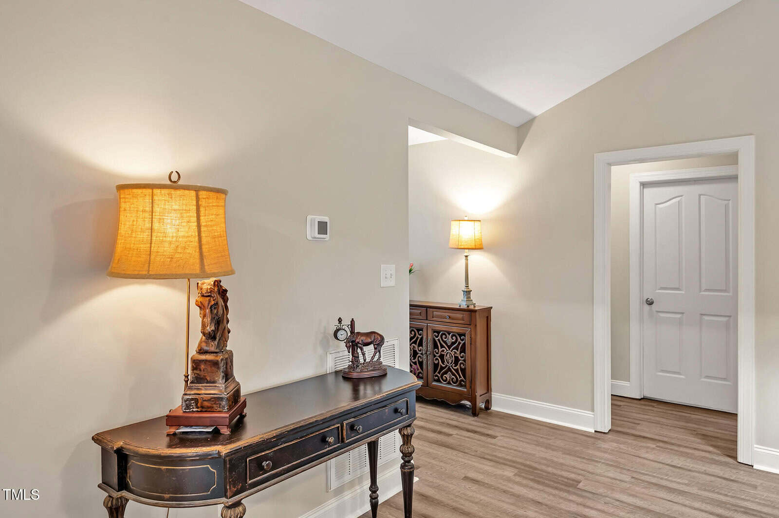 A hallway with light-colored walls and wood flooring features two tables with decorative lamps and ornaments. A closed white door is on the right, and soft lighting creates a warm atmosphere.