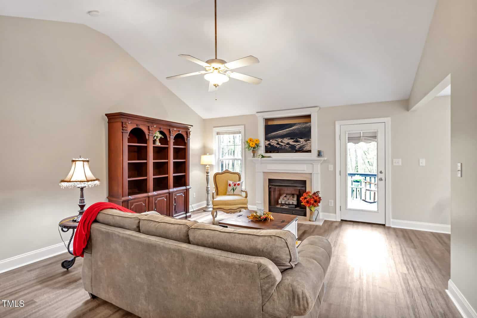Living room with beige walls, vaulted ceiling, ceiling fan, gray sofa, armchair, fireplace, wooden cabinet, lamp, coffee table, and door to outdoor area. A painting and vase of flowers are on the mantel. Light wood flooring.