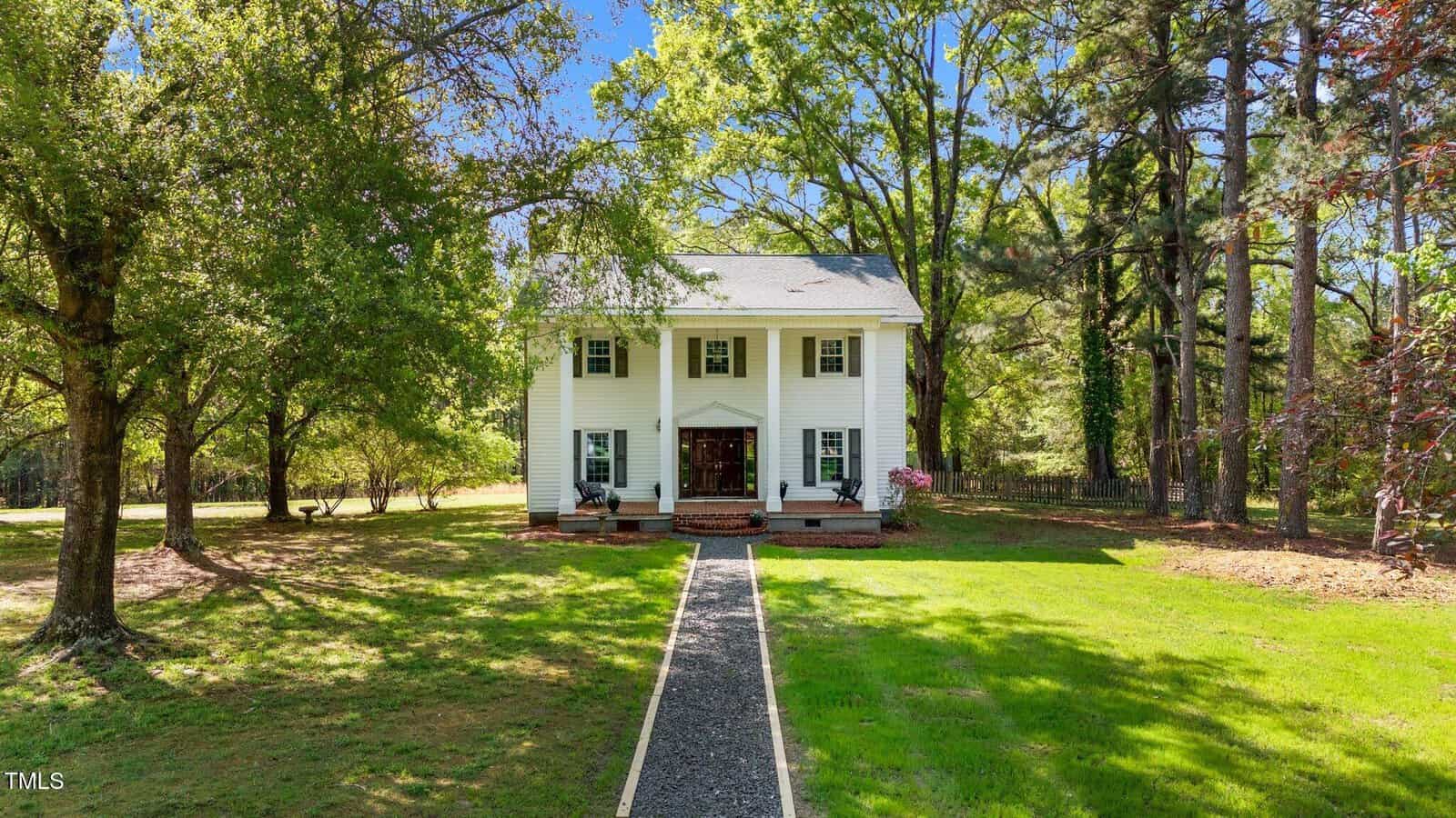 A white two-story house with black shutters and a covered front porch sits on green grass at 1188 Turner Road in Creedmoor, surrounded by trees and a gravel walkway leading up to the entrance on a sunny day.