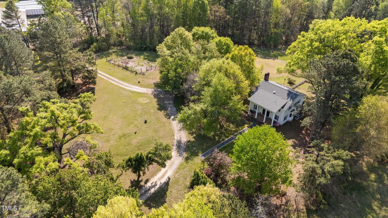 Aerial view of a white house surrounded by trees, with a curved gravel driveway and an open grassy area. A fenced garden or paddock is visible in the background, near the edge of a wooded area.