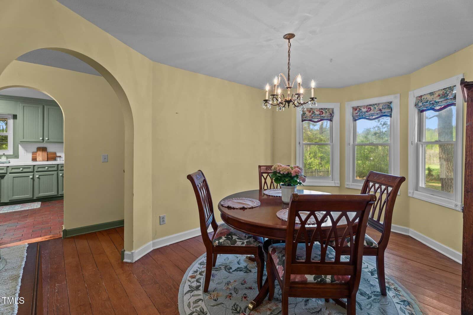A dining room with yellow walls, wood flooring, a round table with four chairs, a floral rug, and a chandelier. Bay windows with valances overlook greenery. An arched doorway leads to a green kitchen.