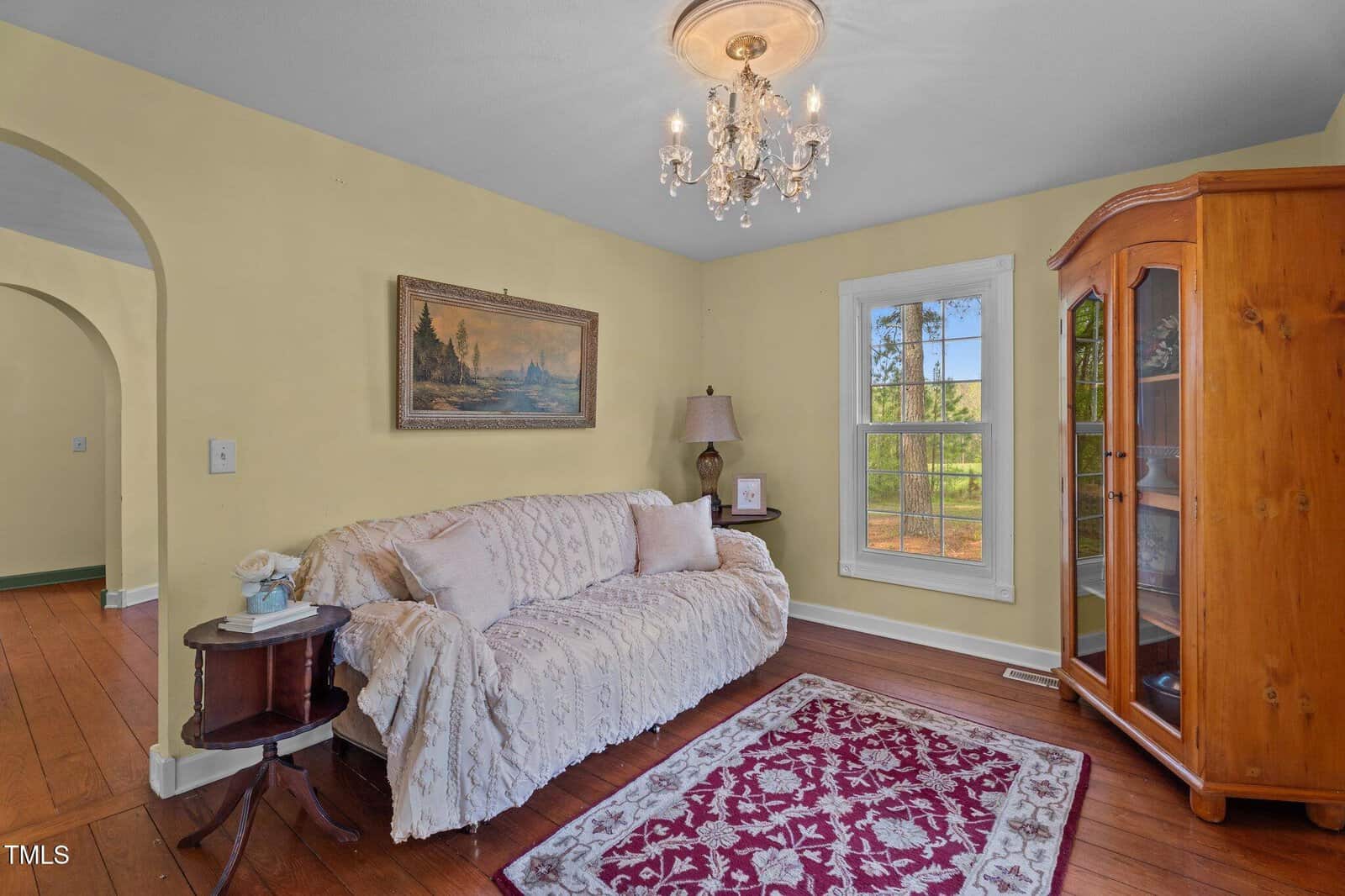 A cozy living room with yellow walls, wood floors, a white couch covered with a lace throw, a red patterned rug, a wooden display cabinet, a window, a lamp, a side table, and a ceiling chandelier.