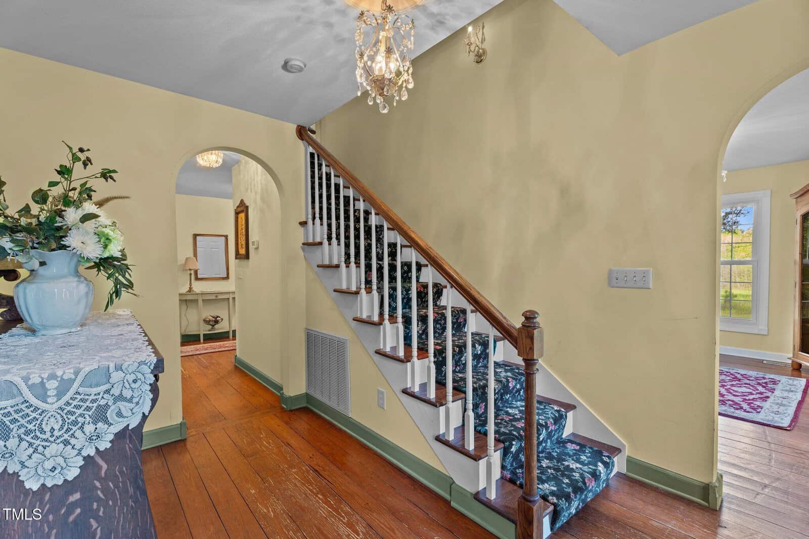 A staircase with a dark wood handrail and patterned carpet runs along a yellow wall. Hardwood floors, a chandelier, arched doorways, and furniture with floral decor are visible in the entryway and adjoining rooms.