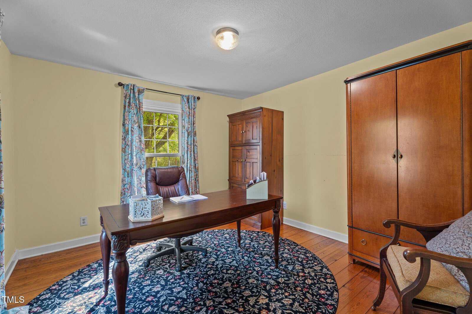 A home office with a wooden desk, chair, and two cabinets. A window with floral curtains lets in natural light. The room has yellow walls, wooden floors, a round rug, and a ceiling light fixture.
