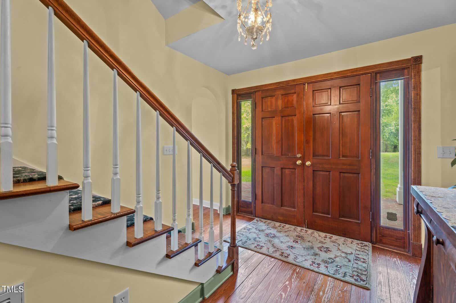 A wooden staircase with white balusters is next to a large double front door made of dark wood. A patterned rug lays on the floor, and natural light enters through glass panels beside the door.