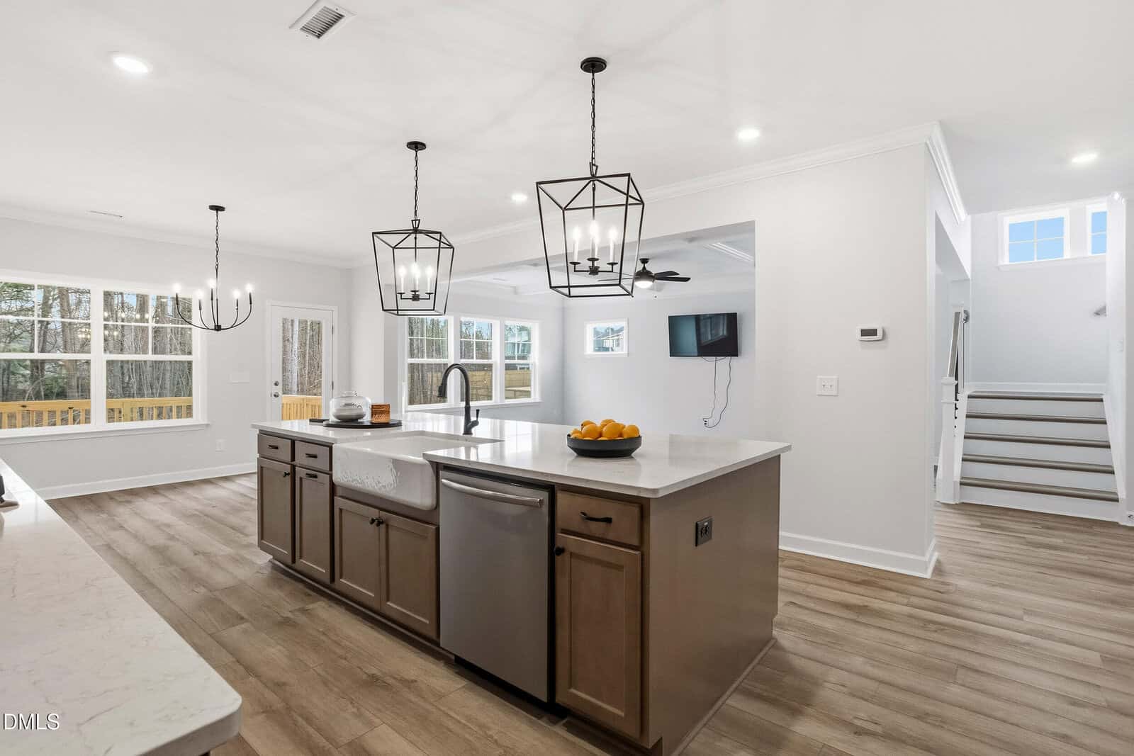 Modern kitchen with a large island, farmhouse sink, pendant lights, stainless steel appliances, and wooden floors. Open layout with a view of the living area, dining space, and staircase. Large windows let in natural light.