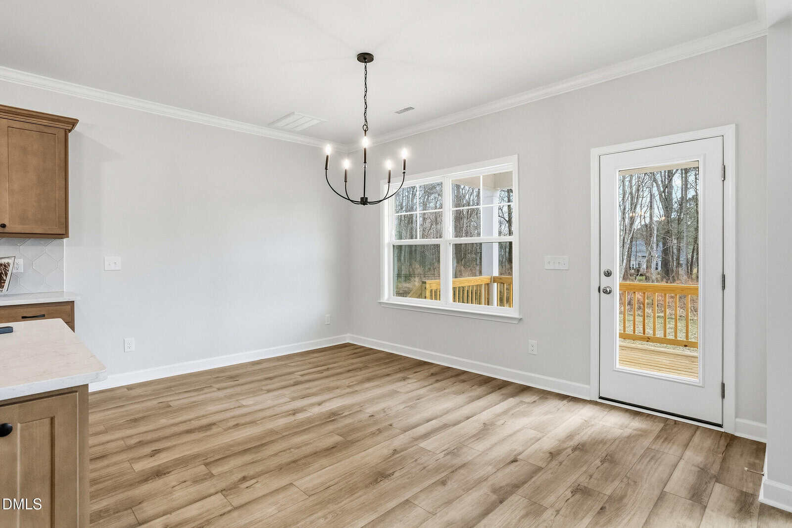 A bright, empty dining area features wood-look flooring, a black modern chandelier, large windows, and a glass door leading to a wooden deck outside. Light gray walls and natural light create an open, airy feel.