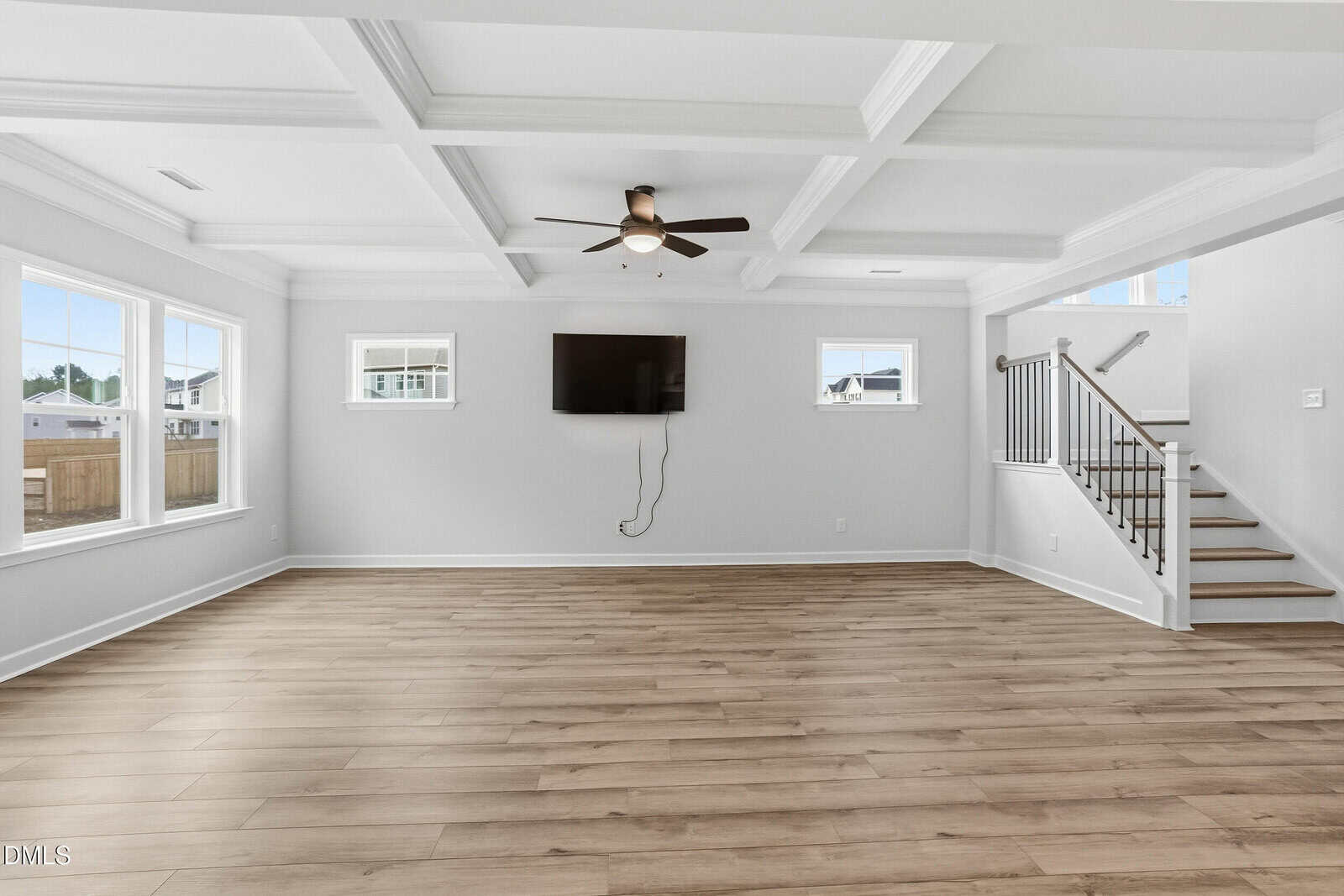 Spacious, empty living room with light wood floors, white walls, large windows, a ceiling fan, wall-mounted TV, and a staircase with white railing leading upstairs.