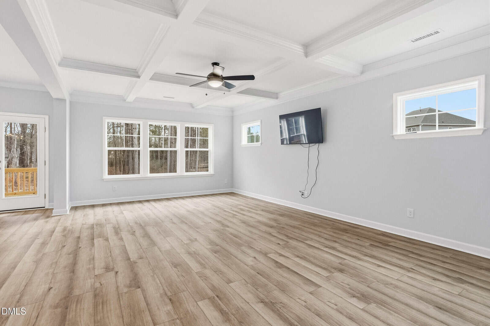 Spacious, empty living room with light wood floors, white walls, large windows, a ceiling fan, tray ceiling, a wall-mounted TV, and a door leading outside. Natural light fills the room.