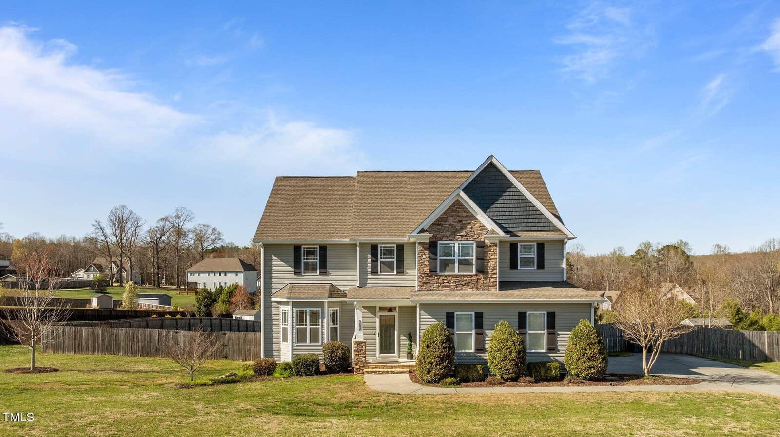 Nestled at 1036 Butterfly Circle in Wake Forest, this charming two-story house boasts gray siding with stone accents and a gabled roof. A neat driveway leads to a front yard adorned with shrubs and trees, while the fenced backyard offers scenic views of neighboring homes against a clear blue sky.
