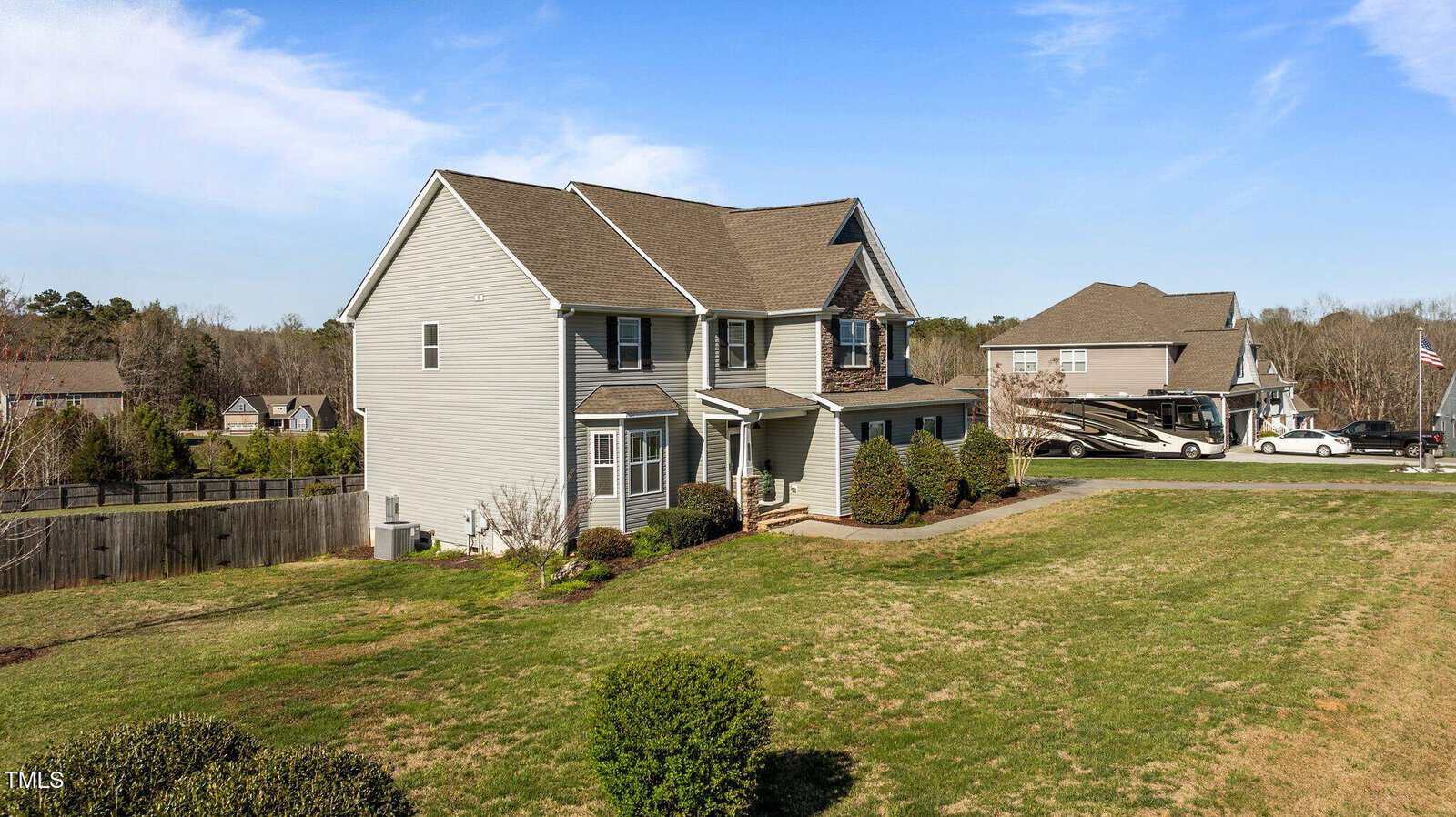 A two-story house with gray siding and a gable roof sits on a large grassy lot. Bushes line the walkway to the front door. Adjacent is a sizable fenced yard. In the background, another house and an RV are visible under a clear blue sky.