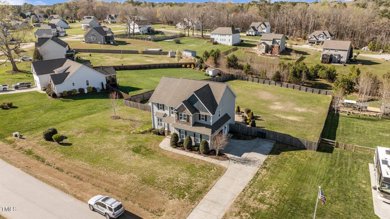 Aerial view of a suburban neighborhood featuring a two-story house with a gray roof and driveway. The house is surrounded by a large, well-maintained lawn and enclosed by a wooden fence. Other houses and green lawns are visible in the background.