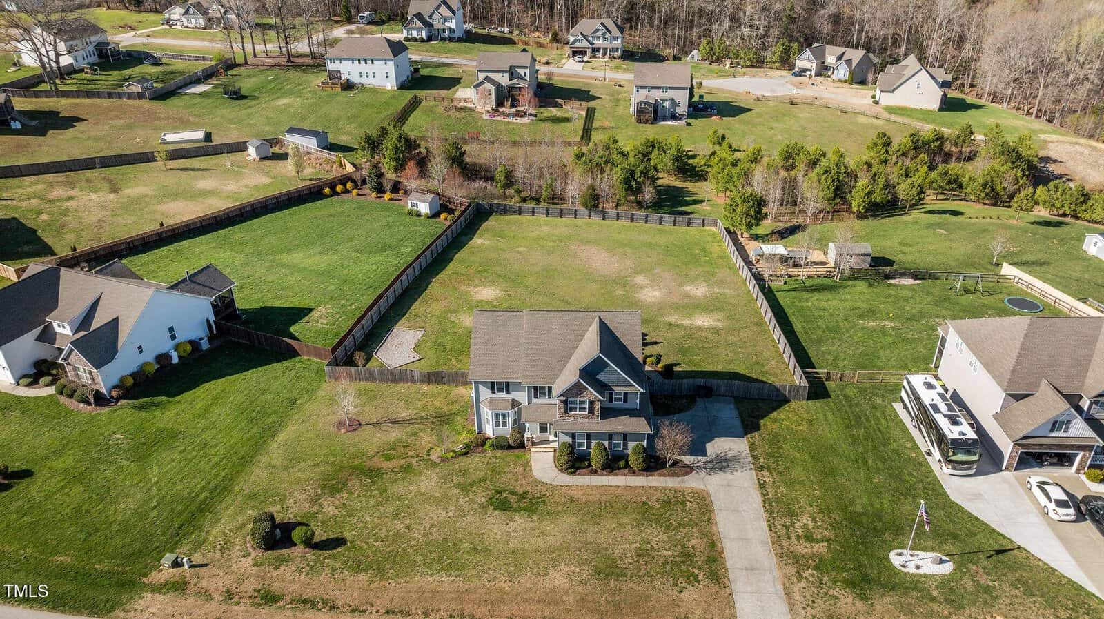 Aerial view of a suburban neighborhood featuring multiple houses with yards. A central house has a large front and back lawn, a driveway, and is surrounded by a fence. Other houses are spread out with trees and open spaces in between.