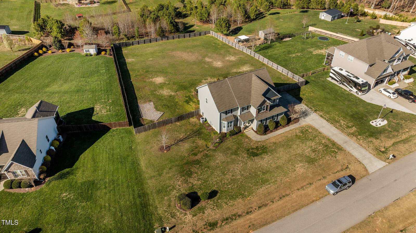 Aerial view of a suburban area featuring a large two-story house with a gray roof, surrounded by fenced green lawns. Adjacent properties also have spacious yards. A car is parked on the street next to the driveway. Trees and additional buildings are visible.