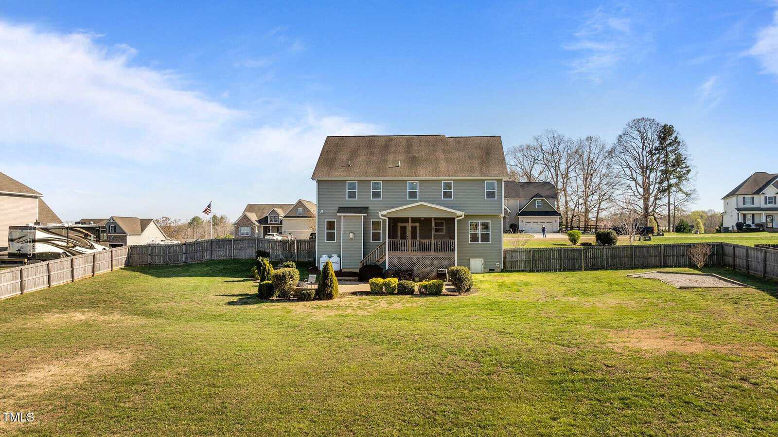 A two-story gray house with a brown roof, featuring a screened porch at the back. It is surrounded by a large fenced yard with trimmed bushes. Neighboring houses and trees are visible under a clear blue sky.