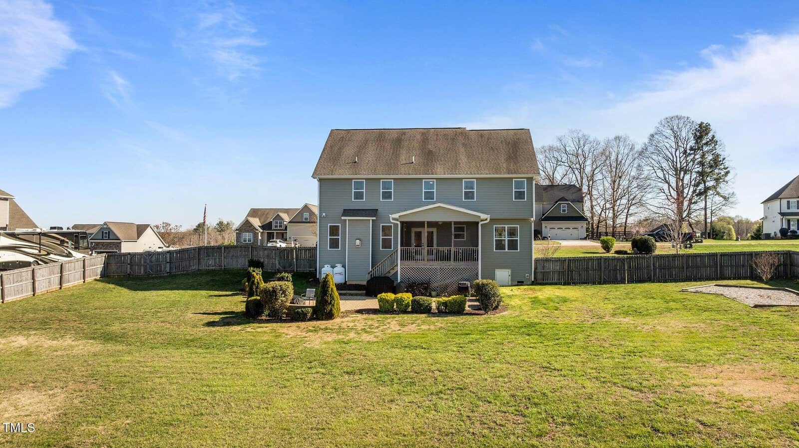 A two-story gray house with a screened porch stands in a spacious backyard. The yard is enclosed by a wooden fence and includes neatly trimmed shrubs. The sky is clear with a few clouds, and neighboring houses are visible in the background.