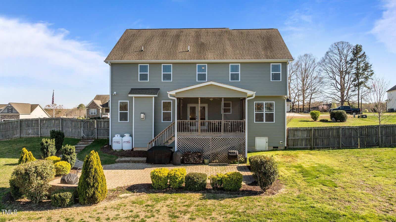 Two-story gray house with a gabled roof, featuring a covered back porch. The yard has a paved patio, bushes, and a fenced perimeter. Propane tanks are visible on the side. The sky is clear and blue.