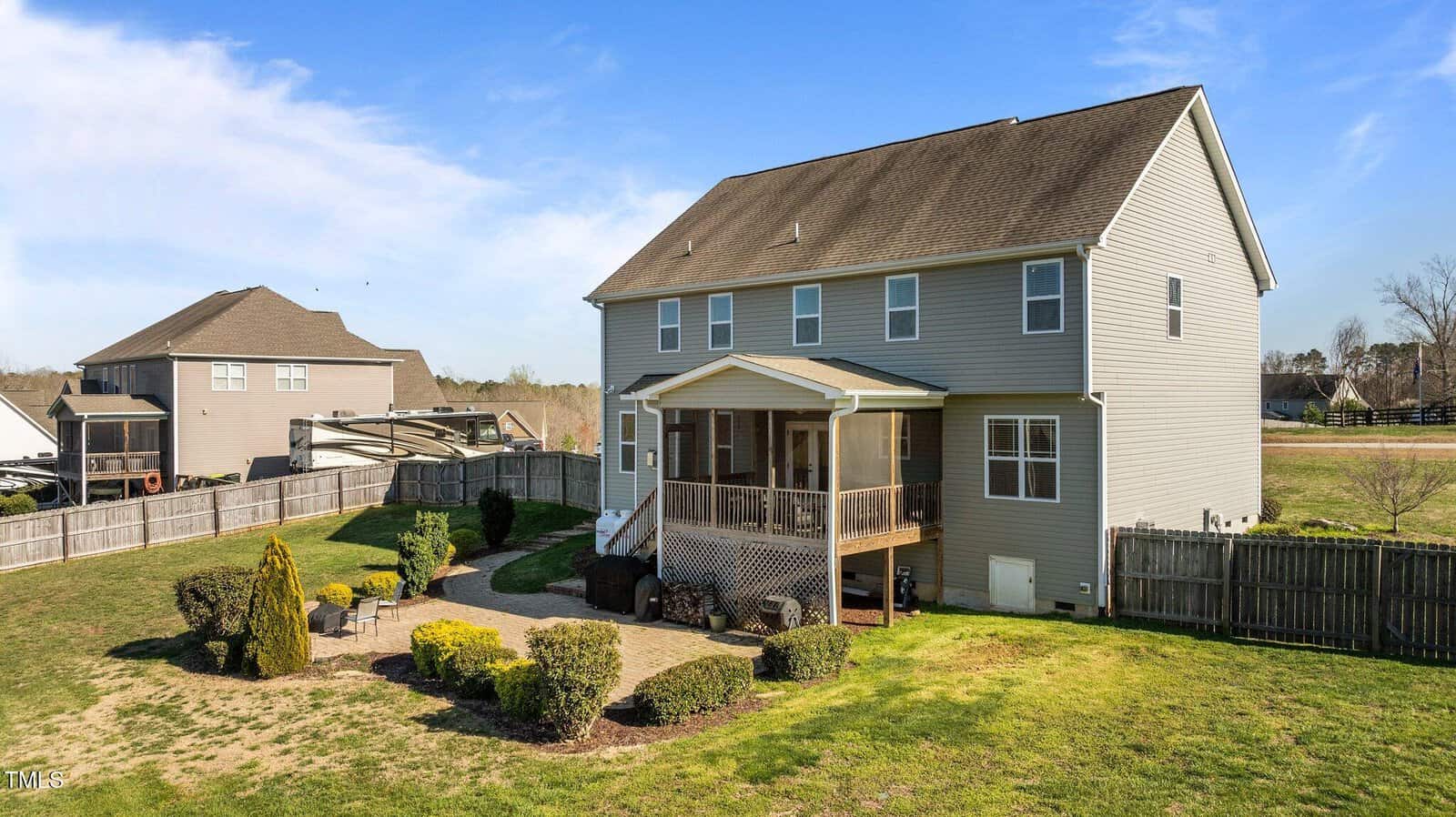 A two-story gray house with a screened porch, fenced backyard, manicured shrubs, a patio seating area, and neighboring homes in the background under a clear blue sky.