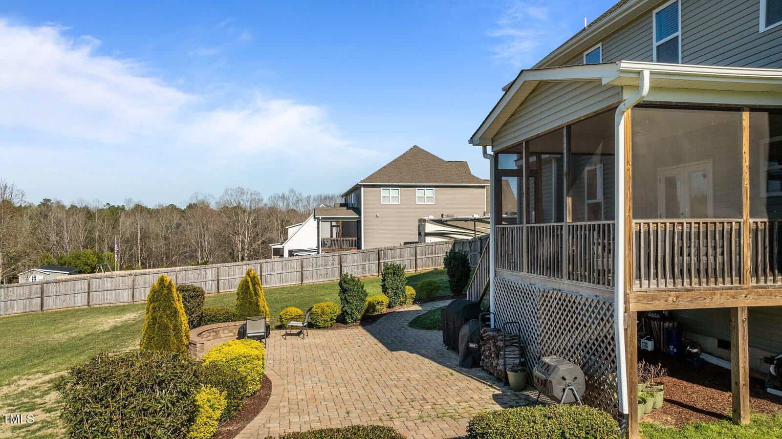 A backyard patio with pavers, green shrubs, and a screened-in porch attached to a house. Wooden fencing and neighboring homes are visible in the background under a blue sky.