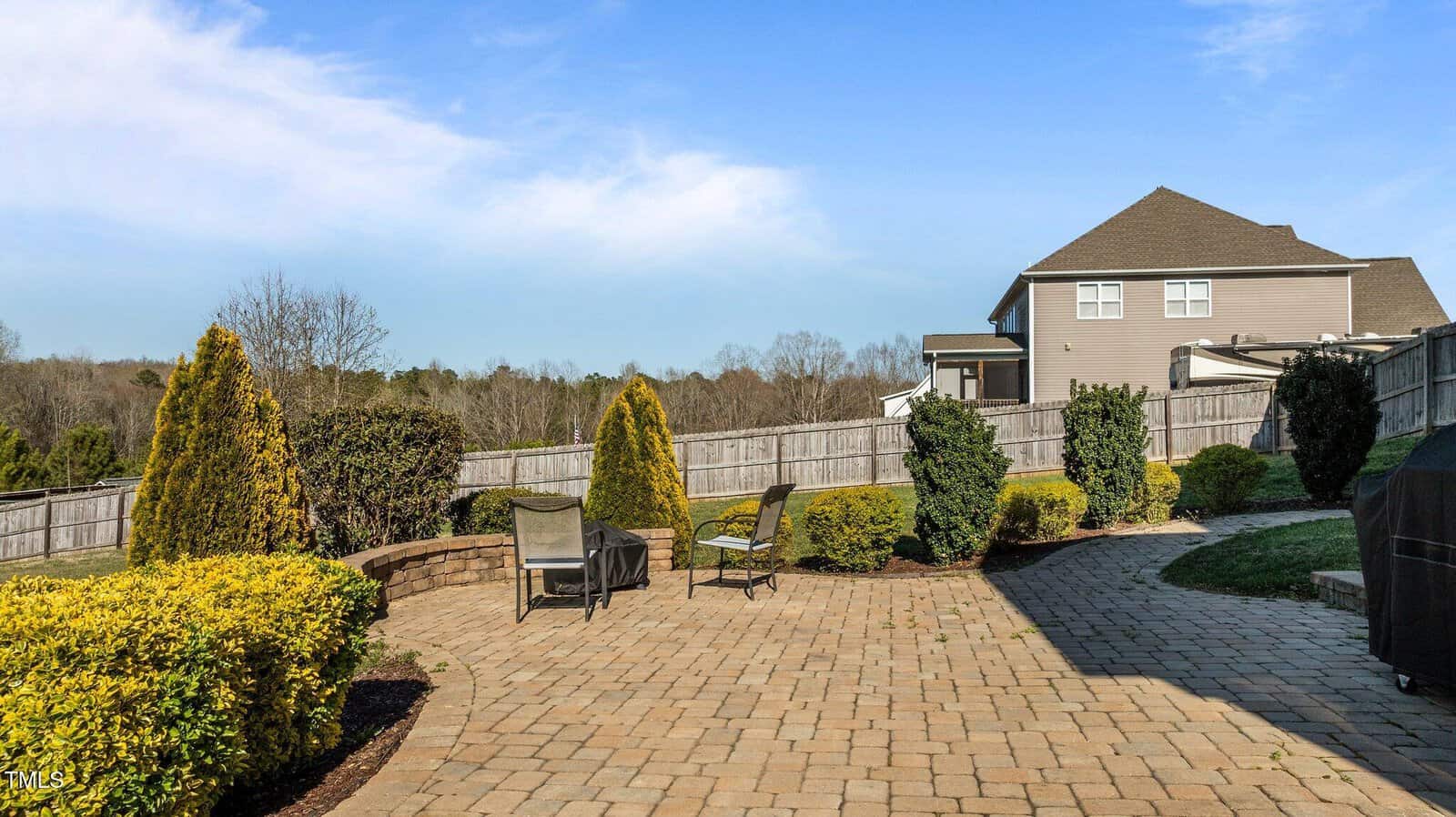 A backyard patio with two chairs and a small table sits on stone pavers, surrounded by shrubs, small trees, and a wooden fence. A house and grill are visible under a blue sky.