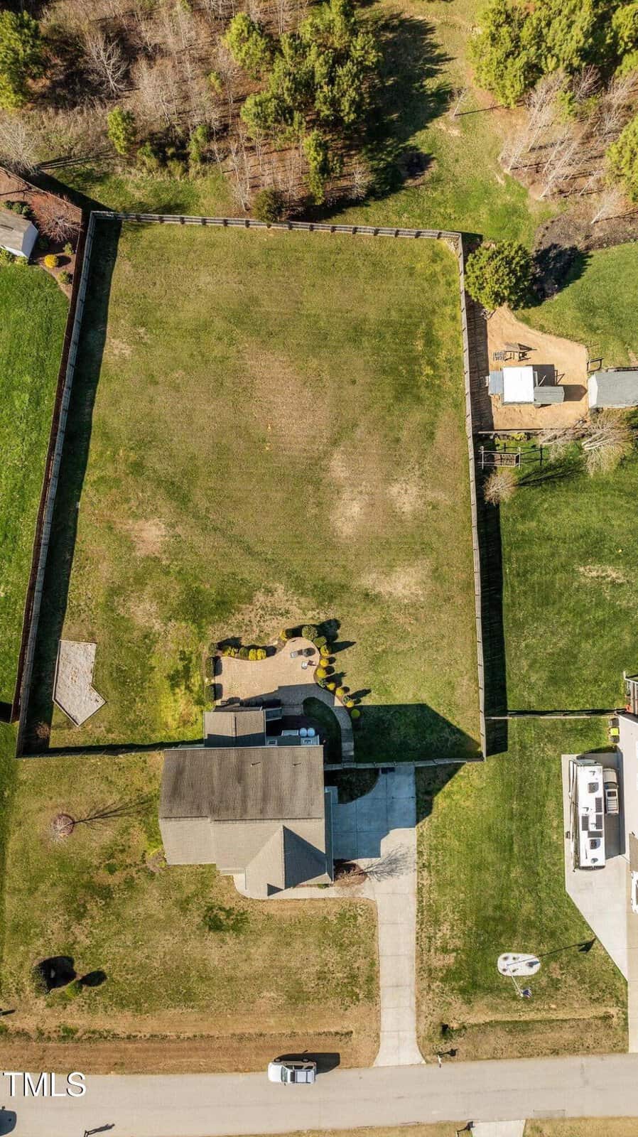 Aerial view of a suburban house with a large, fenced backyard. There is a driveway leading to the house and a patio area adjacent to it. Trees and grass surround the property, with neighboring homes and an RV visible nearby.