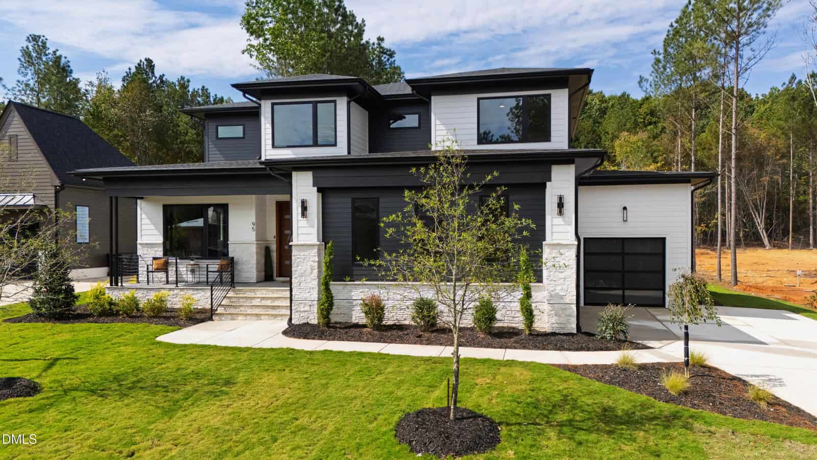 Modern two-story house at 95 Cherry Bark Drive, Youngsville, featuring a black and white exterior, large windows, front porch with seating, attached single-car garage, landscaped yard with young trees and neat grass. Forested backdrop completes the scene.