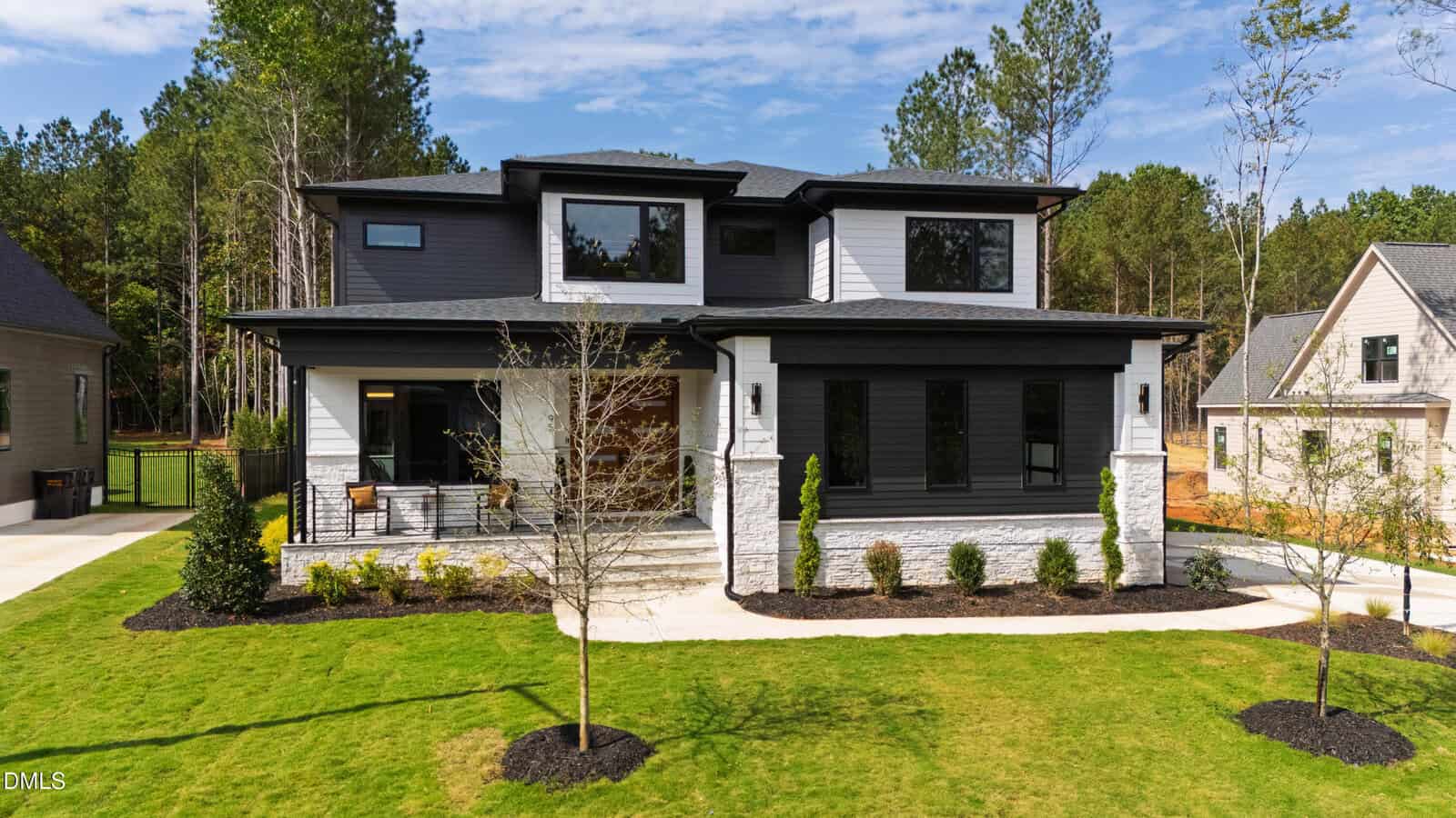 Modern two-story house with black and white exterior, large windows, and a covered front porch. It is surrounded by a neatly manicured lawn, young trees, and sits in a suburban neighborhood with wooded surroundings.