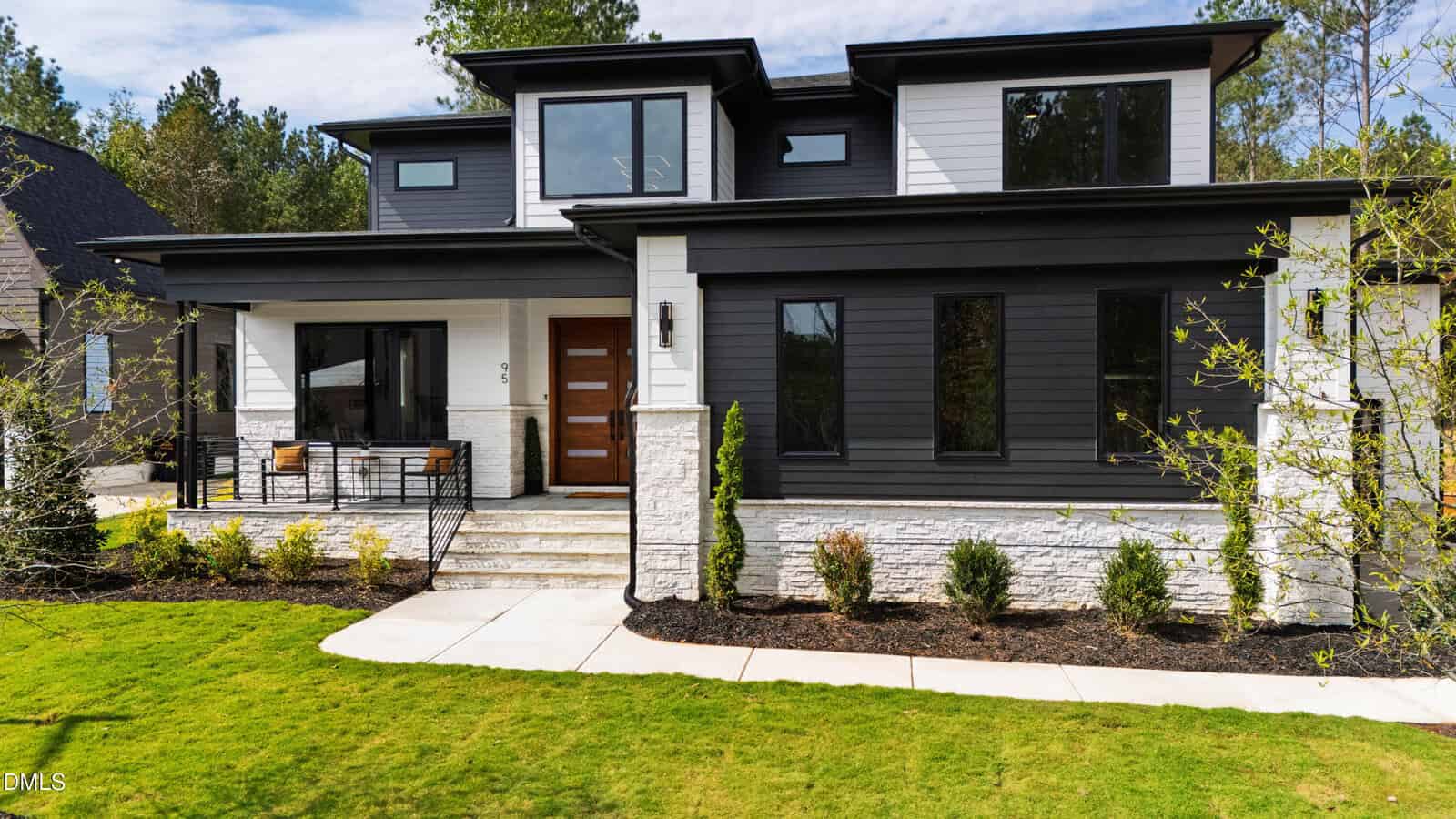 A modern two-story house with black and white exterior, large windows, a wooden front door, a small porch with chairs, landscaped yard, and green lawn under a blue sky.