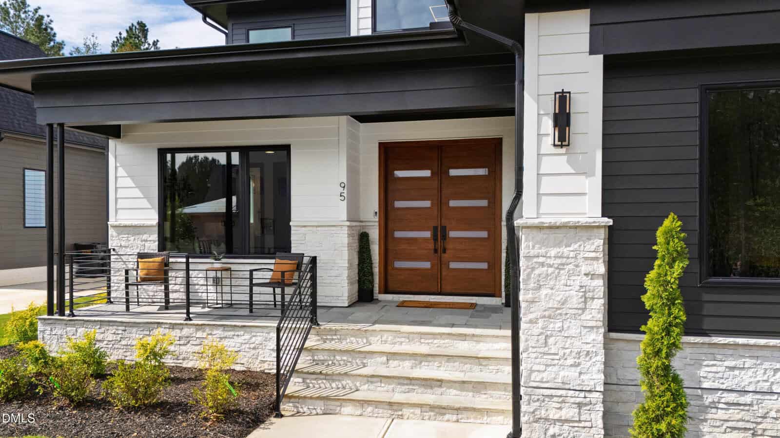 Modern house exterior with dark and white siding, stone accents, large double wooden front doors, a small front porch with chairs and a table, and minimal landscaping.