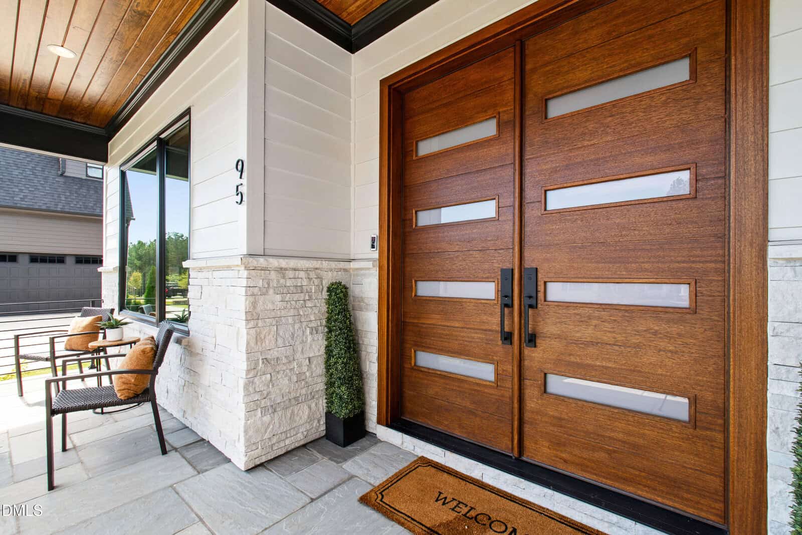 Modern front porch with a large wooden double door featuring horizontal glass panels, white stone and siding exterior, potted topiary, welcome mat, and outdoor seating area with chairs and cushions.